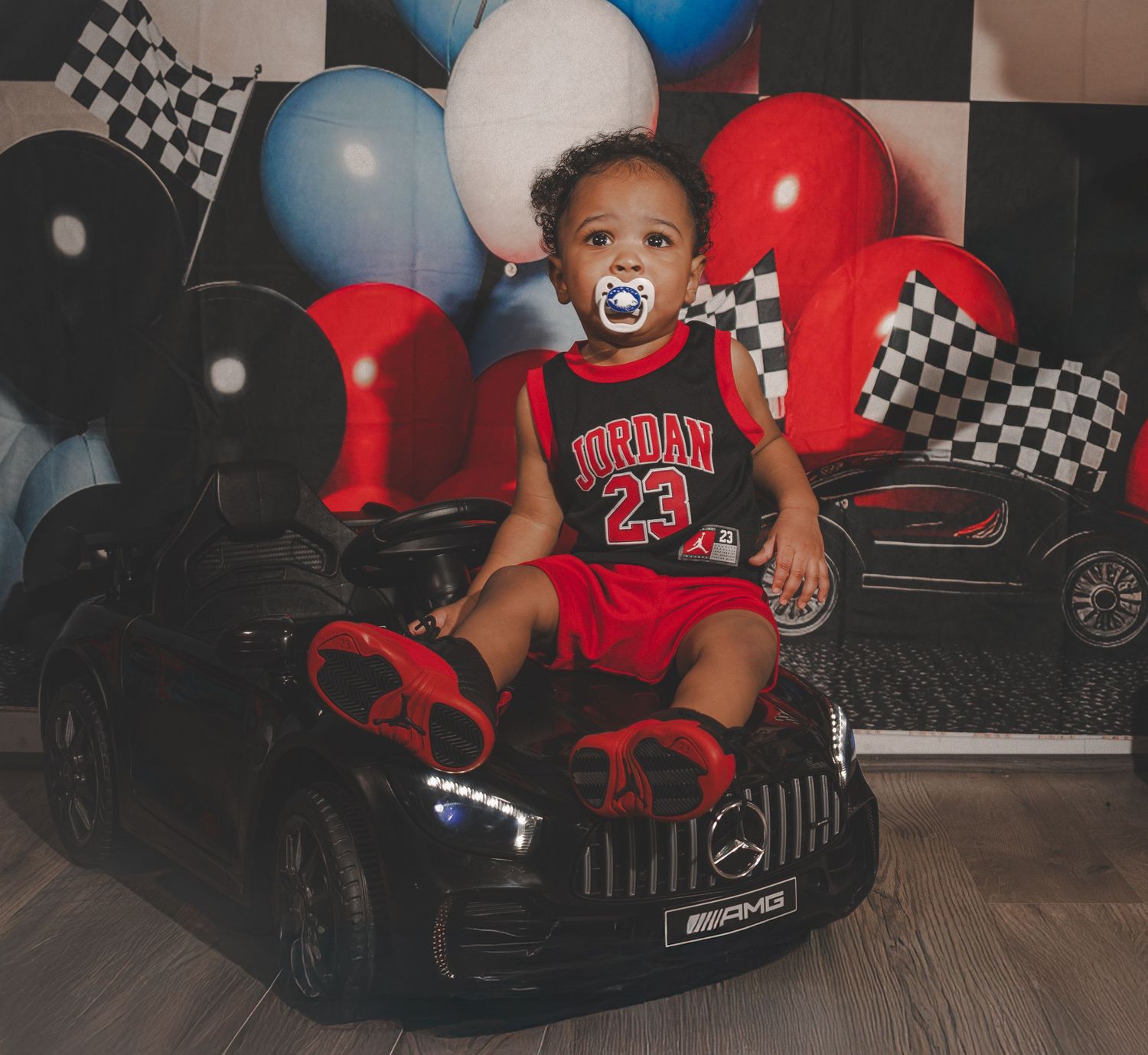 Child playing in a toy electric car with black and white checkered racing flags and red balloons in background.