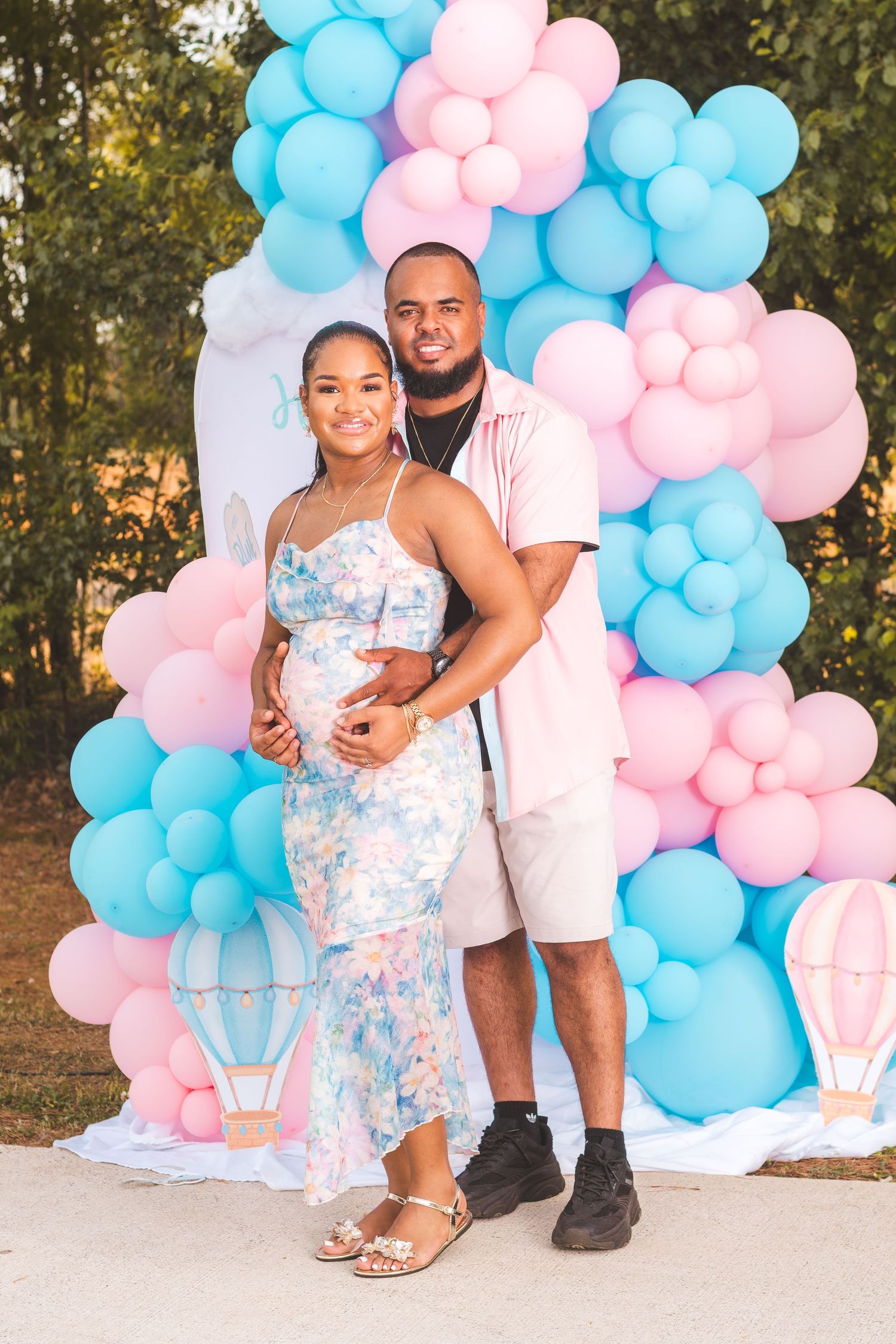 Couple posing at a gender reveal party with pink and blue balloon arch backdrop outdoors.