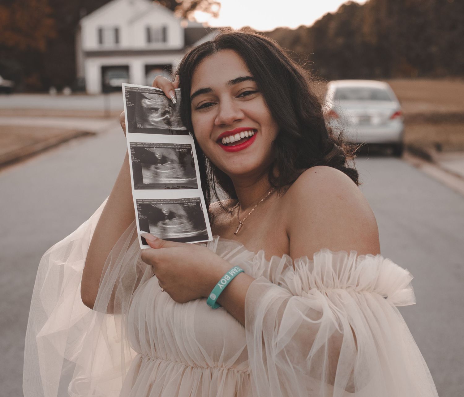 Someone in a white tulle dress holds ultrasound photos while smiling outdoors.