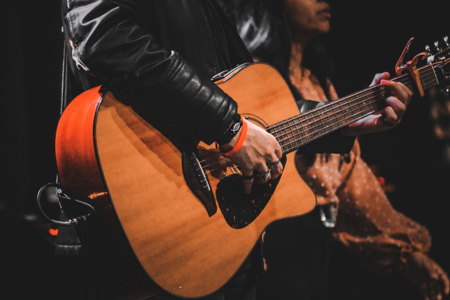 Close-up shot of hands playing an acoustic guitar with leather jacket sleeve visible.