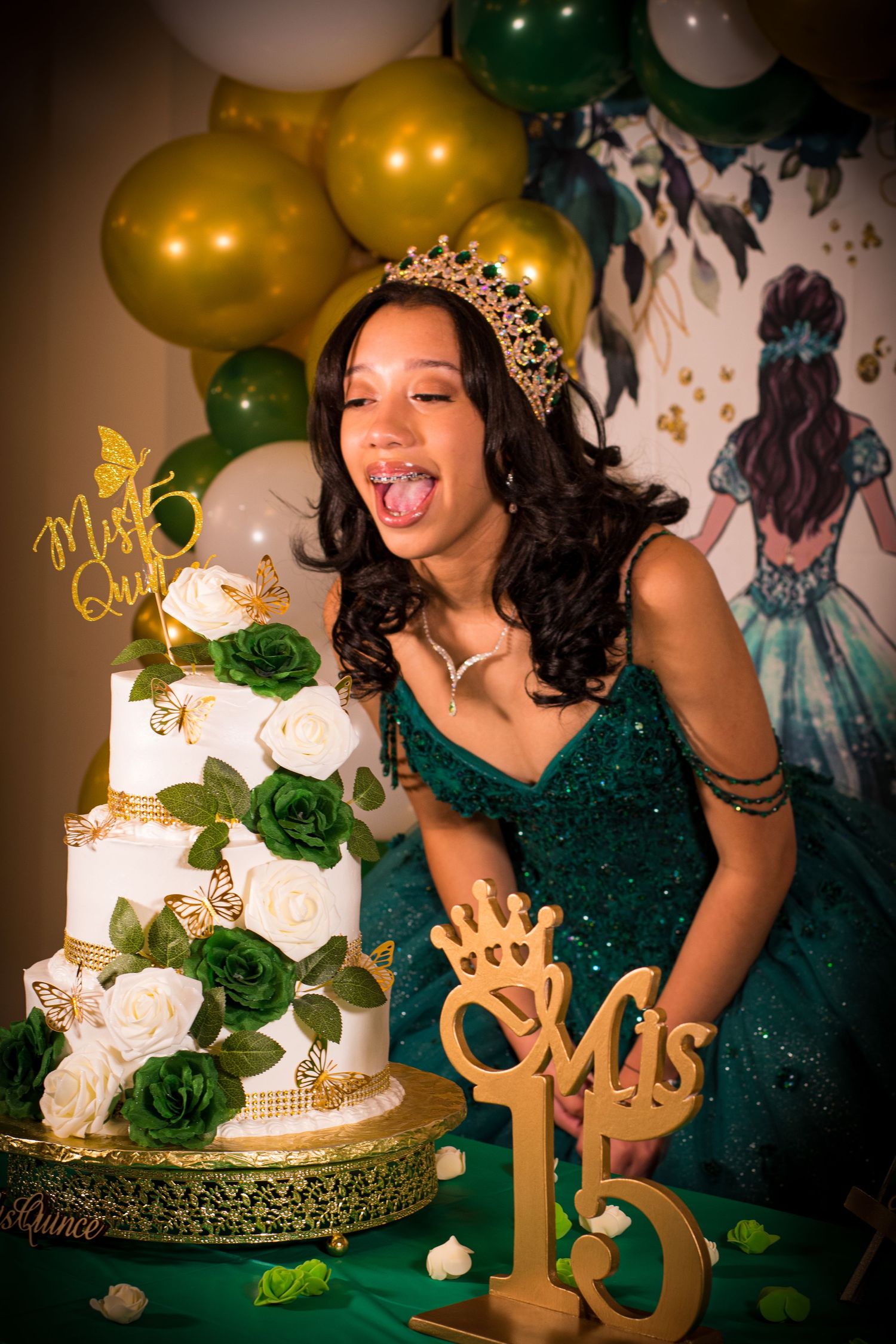 Celebrant with tiara poses excitedly next to an elegant white and green floral cake with gold decorative elements.