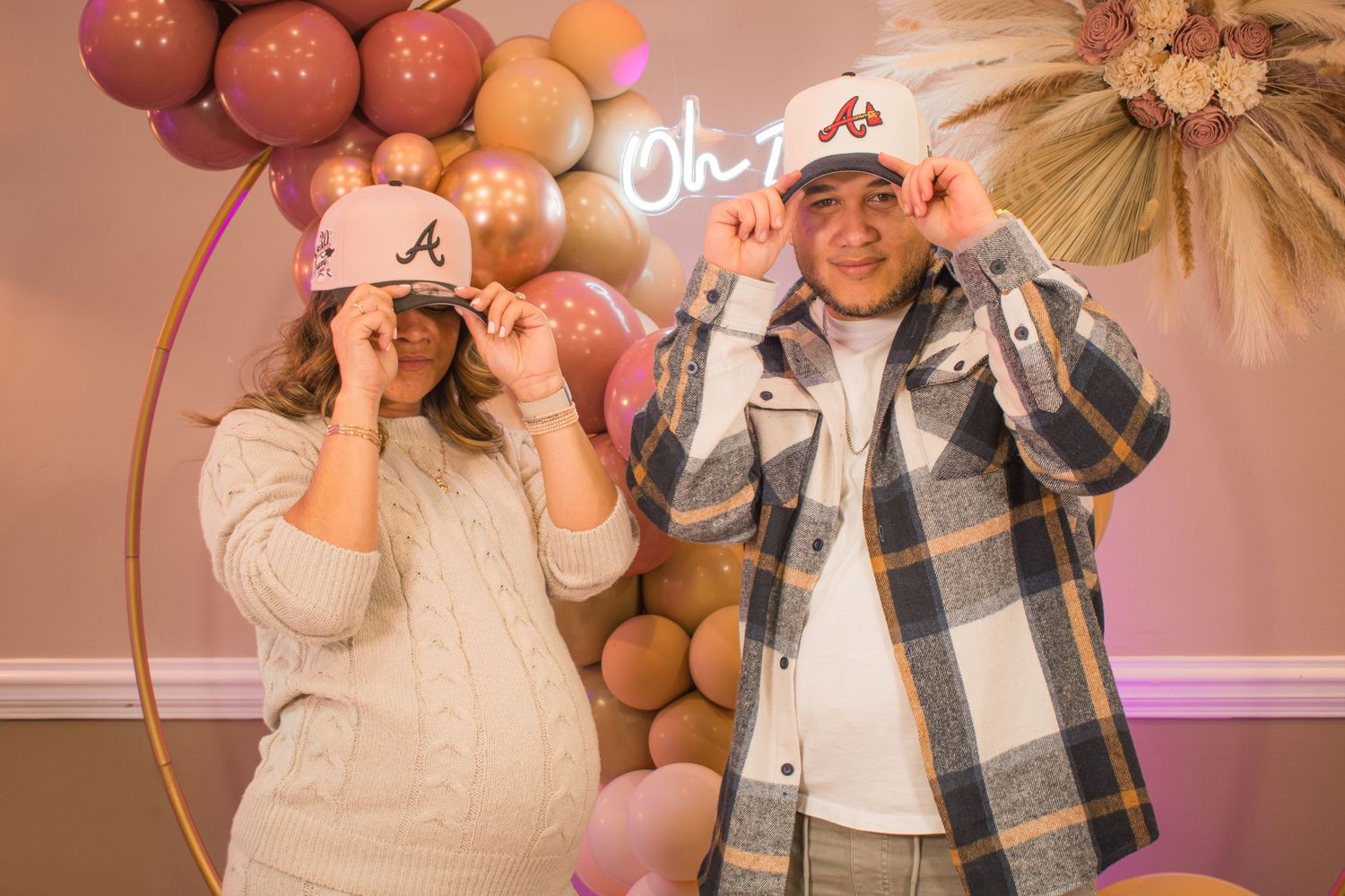 A playful couple wearing matching Braves baseball caps pose against a balloon backdrop.