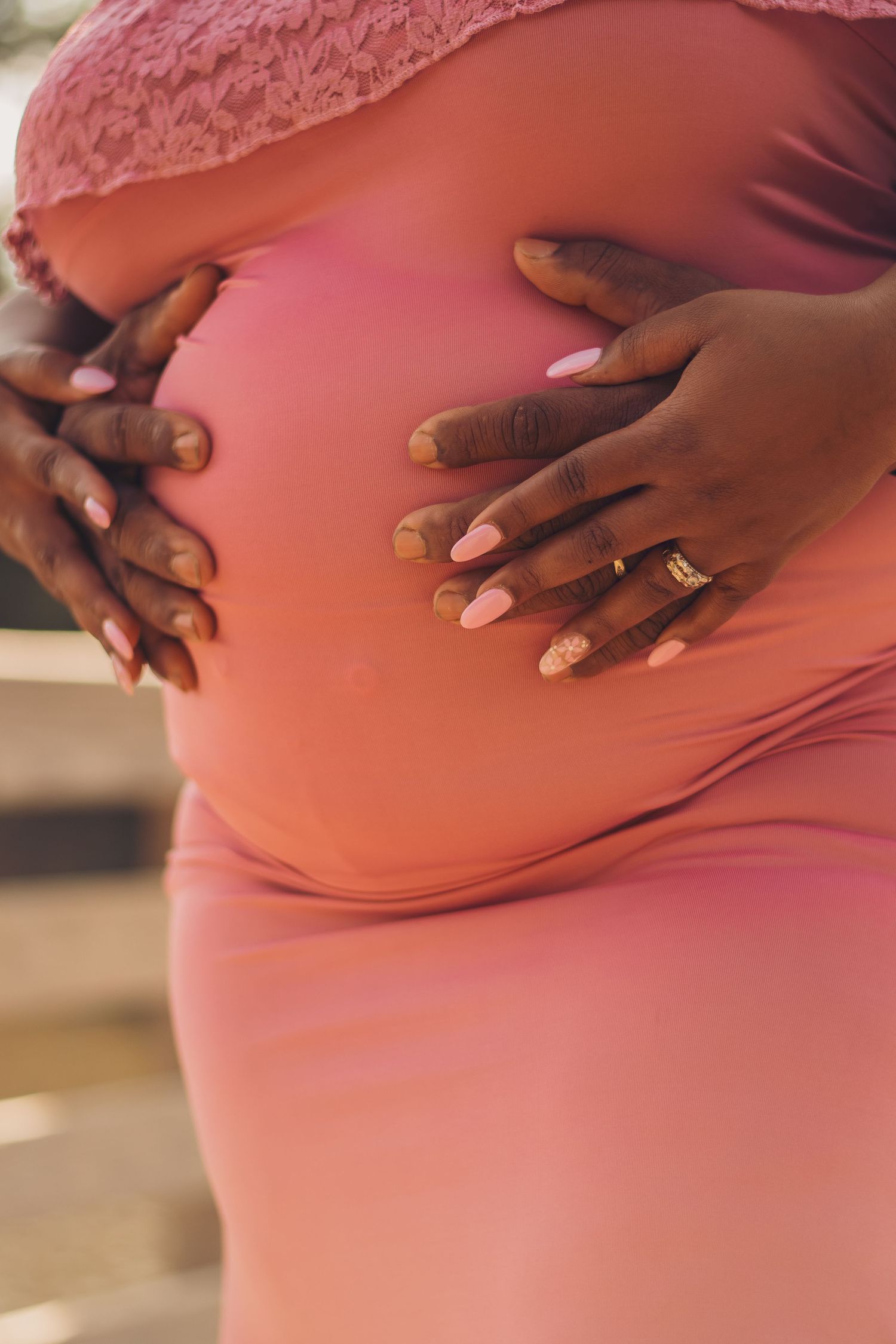 Close-up of hands tenderly cradling pregnant belly in coral maternity dress.