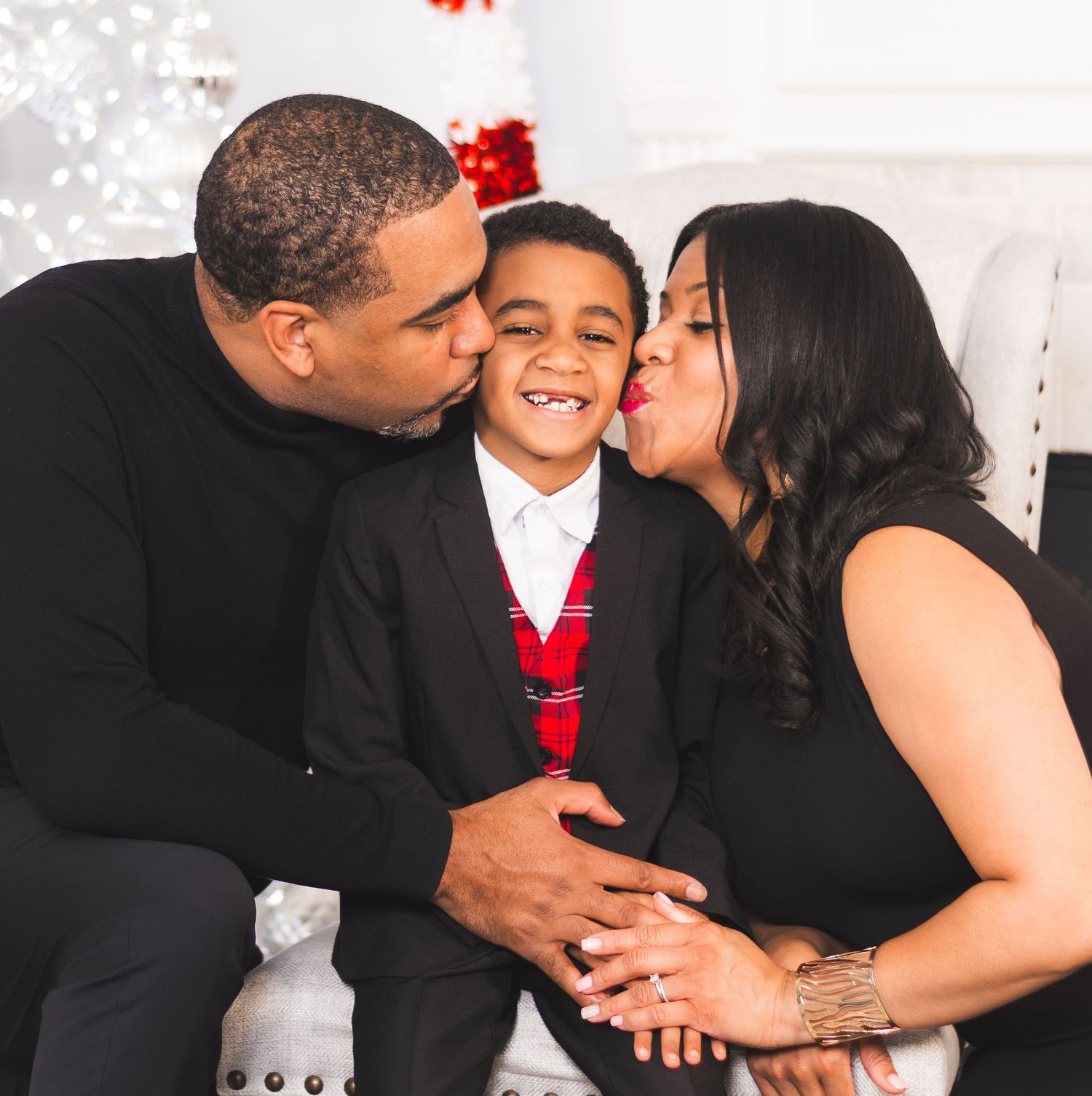 A joyful family Christmas portrait with parents kissing their young son on the cheeks in a white holiday setting.