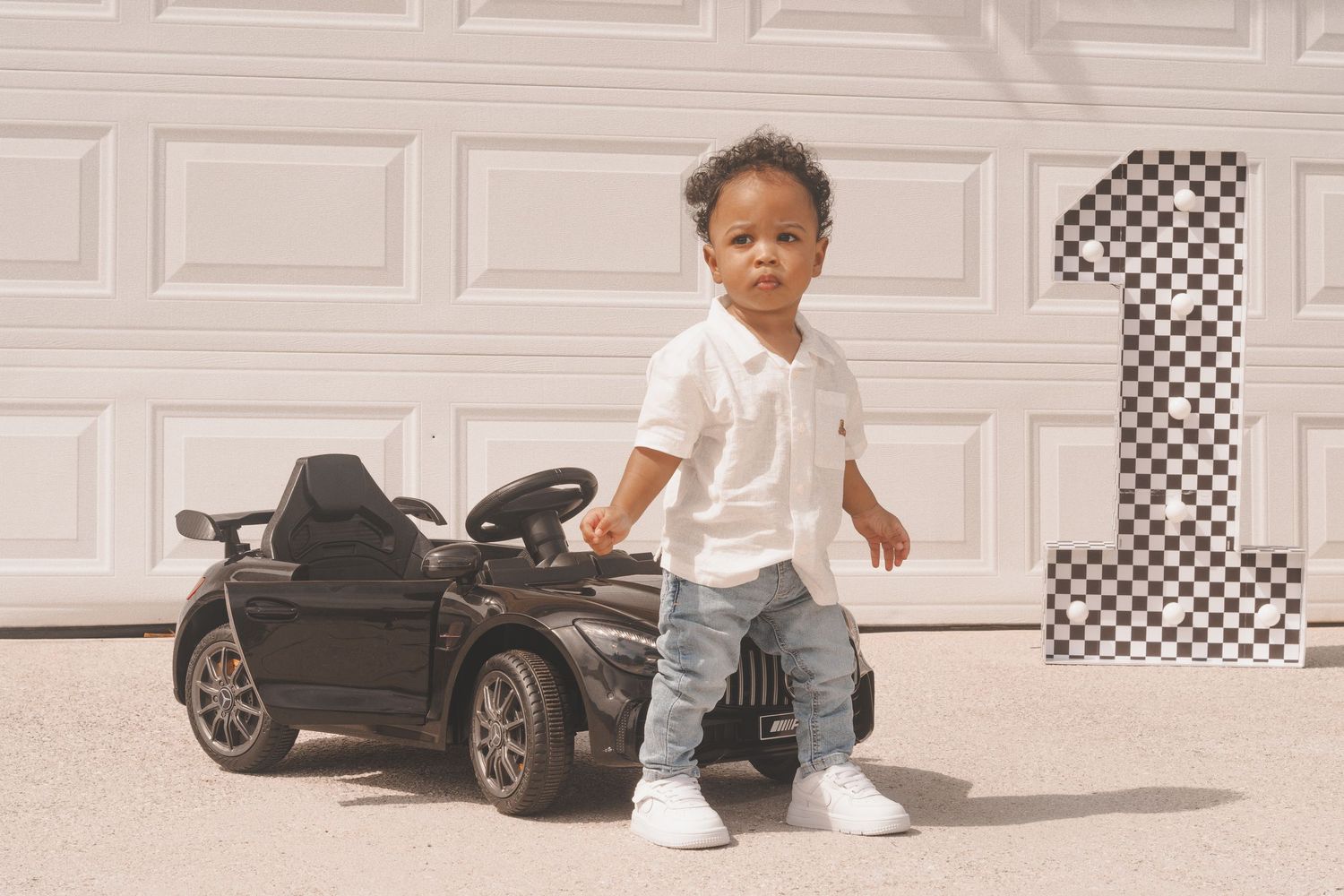 A toddler stands next to a black toy ride-on car with a checkered number one decoration on a white garage door.