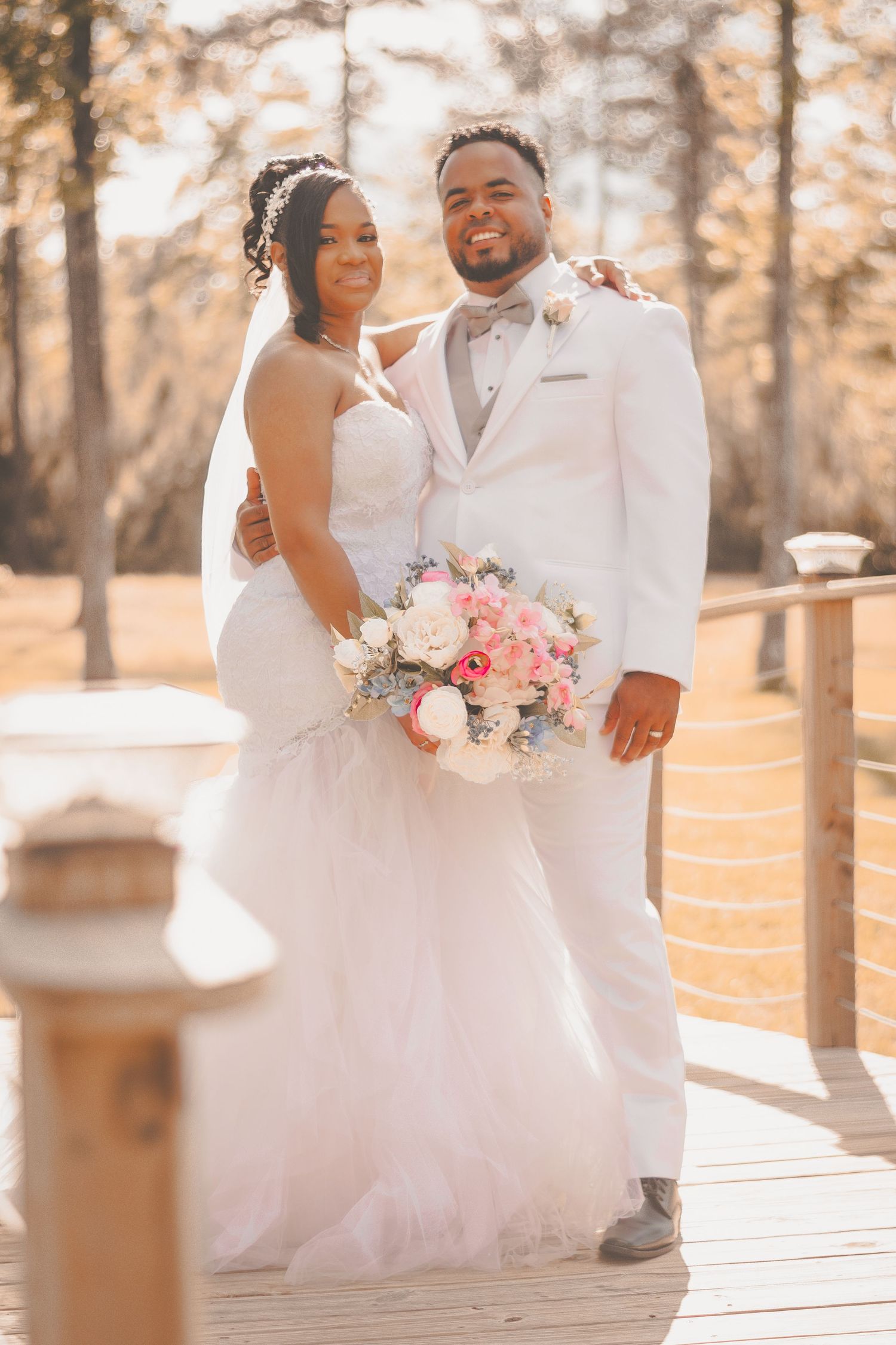 A romantic wedding photo showing a newlywed couple embracing on a wooden deck holding a pink and white bouquet.