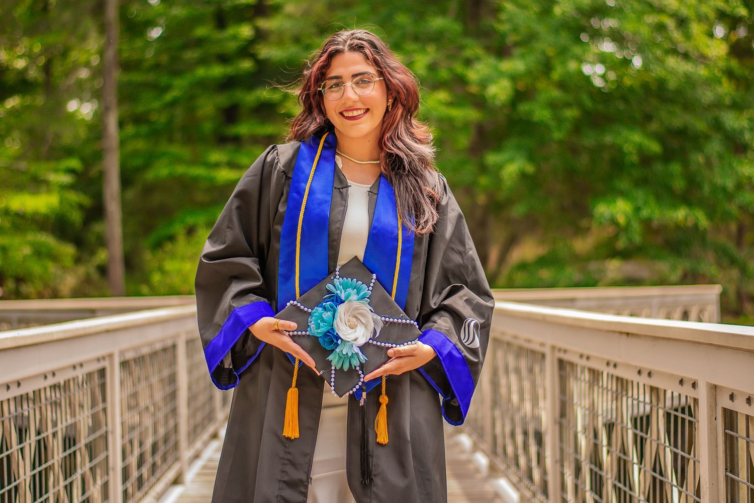 Graduate in cap and gown holding decorated graduation cap on wooden bridge.