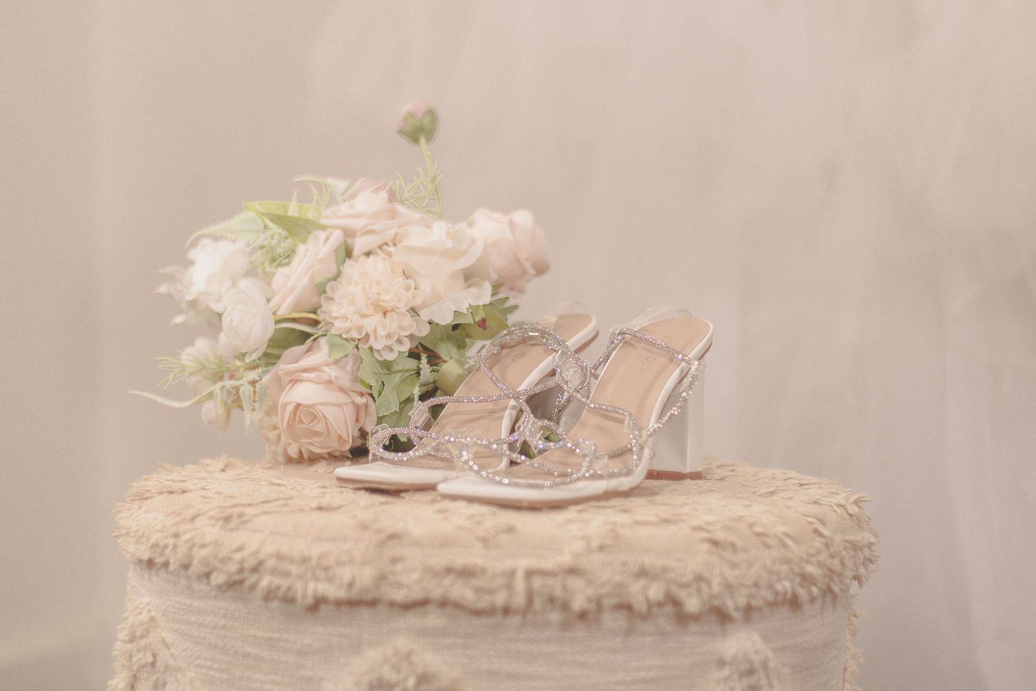 Delicate silver bridal sandals beside pink floral bouquet on light beige textured surface with soft vintage aesthetic.