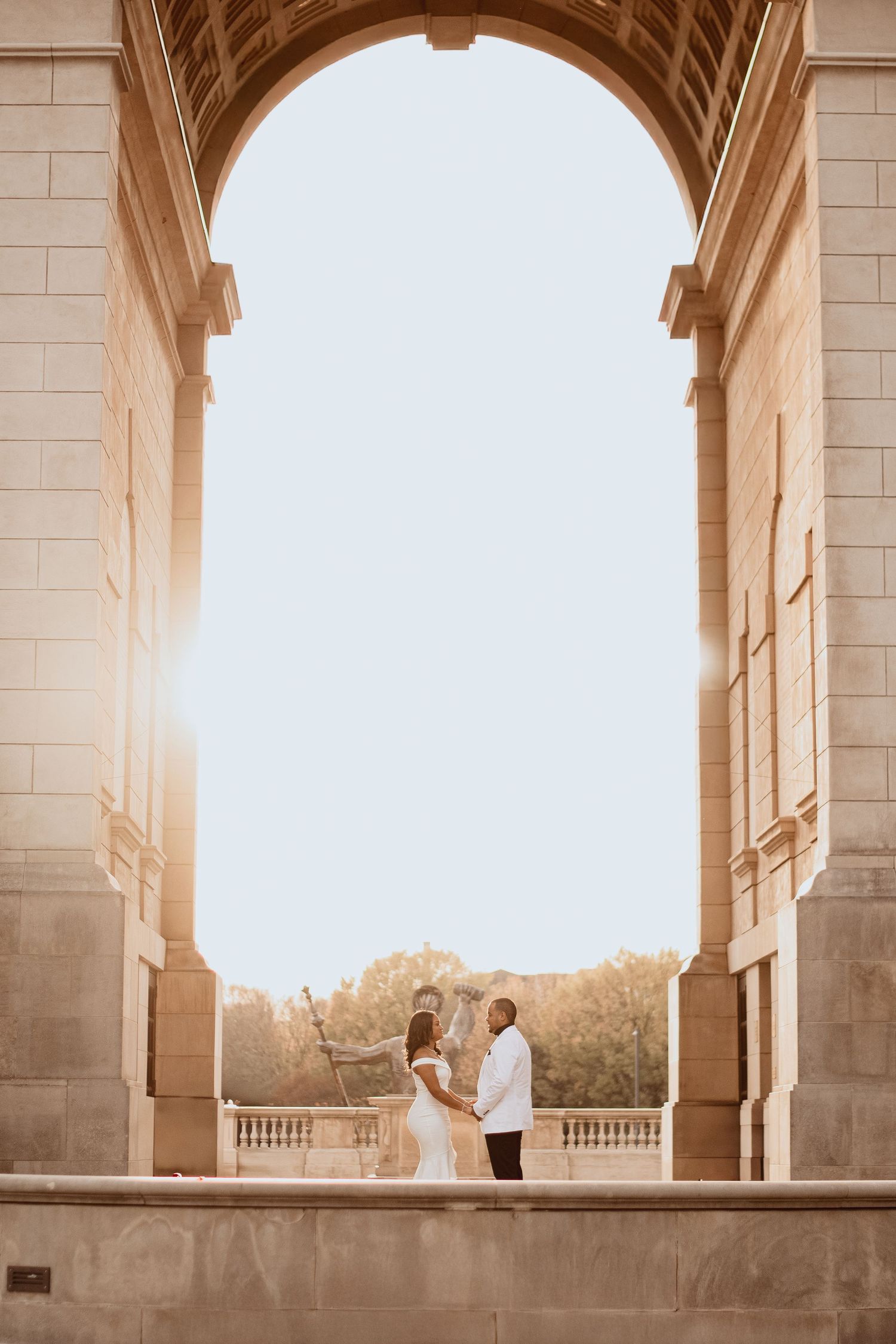 Romantic couple standing in a grand stone archway at sunset, silhouetted against bright sunlight.