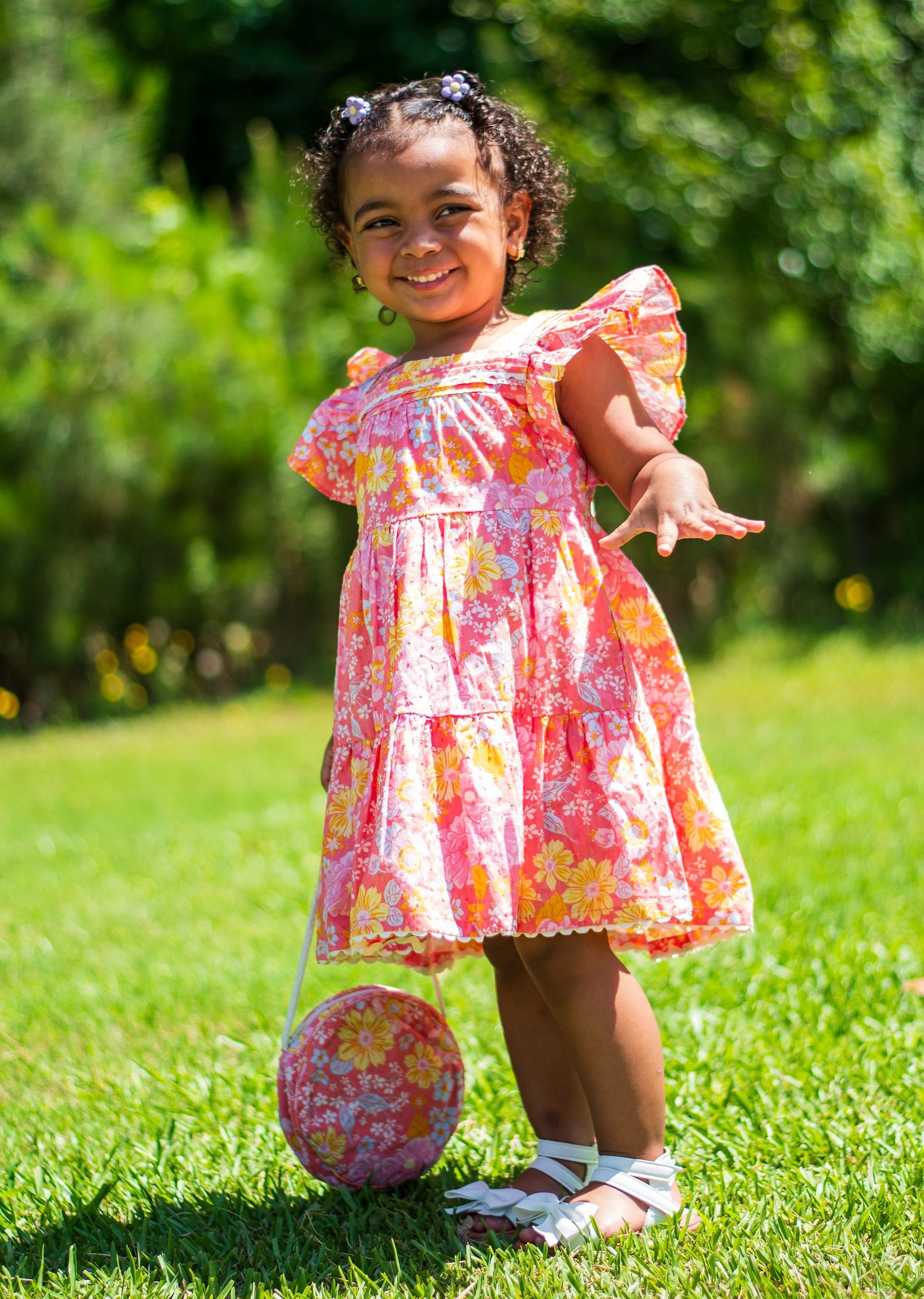 Smiling toddler in pink floral dress posing with matching purse on grass.