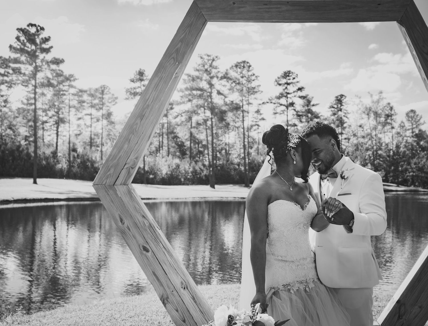 A romantic couple embraces inside a geometric frame beside a serene lake surrounded by tall pine trees in black and white.