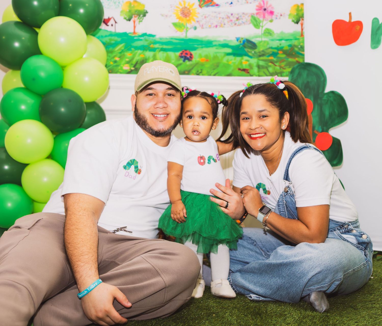 Family of three posing at a Very Hungry Caterpillar first birthday party with green balloon arch.