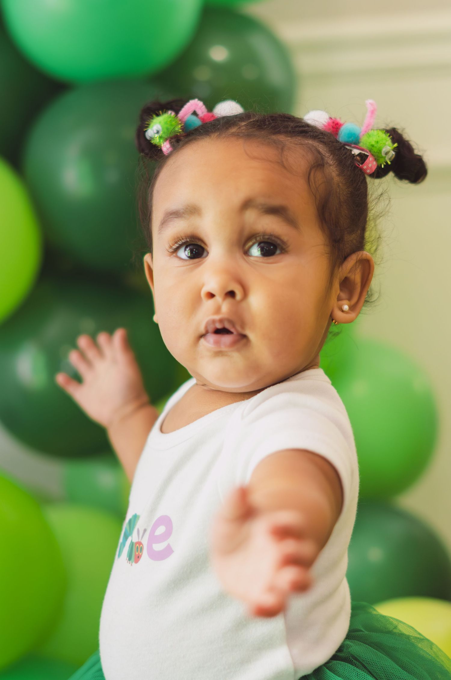 Adorable baby girl in green tutu and colorful hair accessories celebrates first birthday with green balloon backdrop.