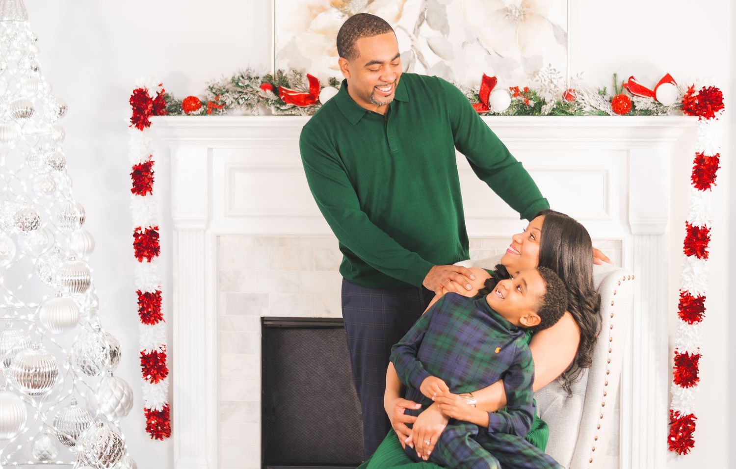 Couple in festive green outfits smiling by a decorated Christmas fireplace mantel.