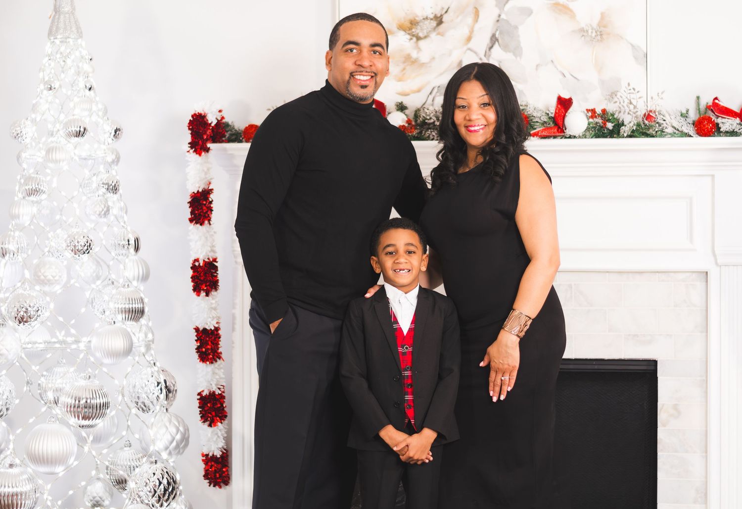 Family of three posing for a Christmas portrait near a white tree and fireplace decorated with red and silver holiday decor.