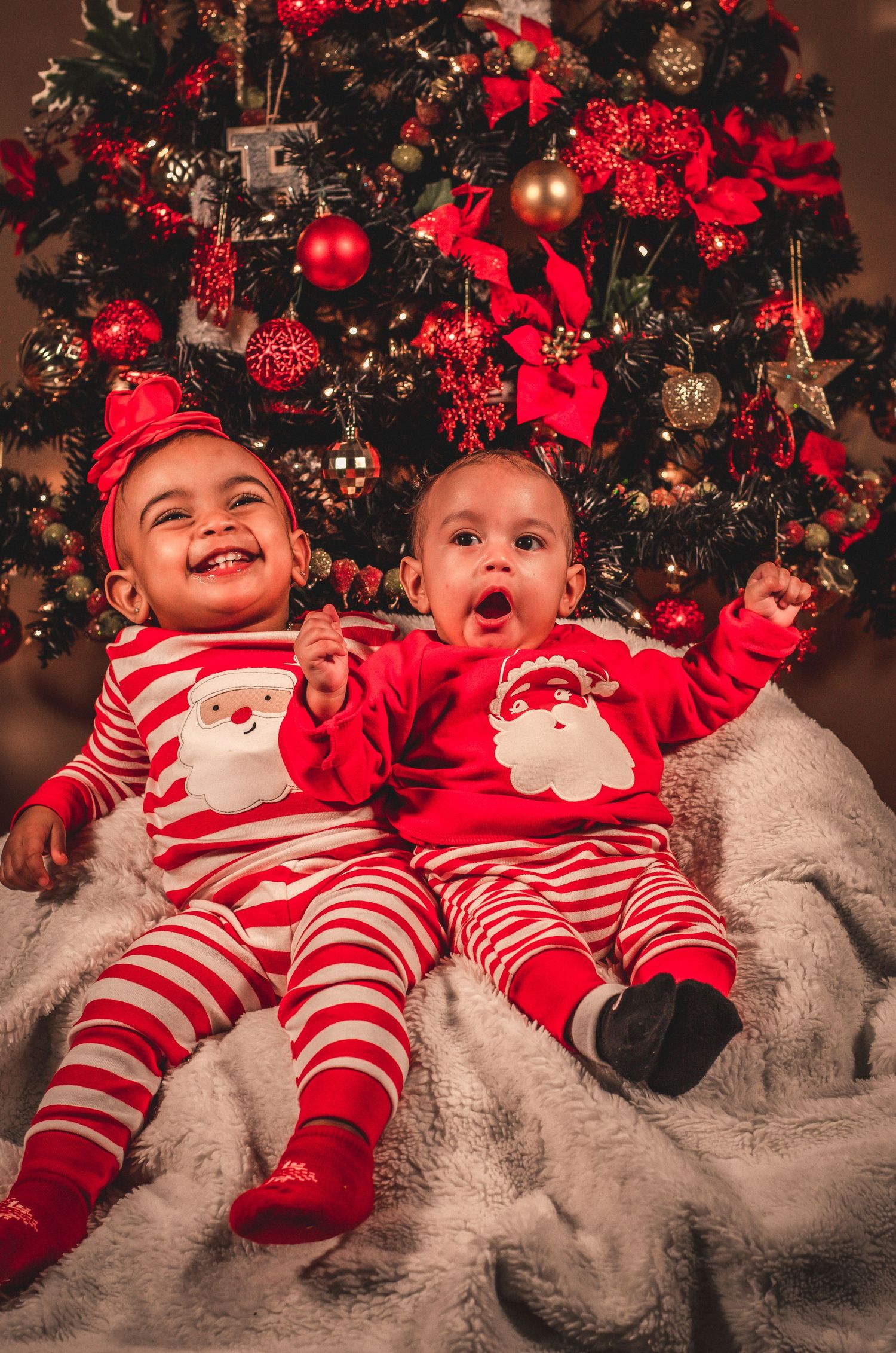 Two babies in matching Santa striped pajamas laughing by Christmas tree.