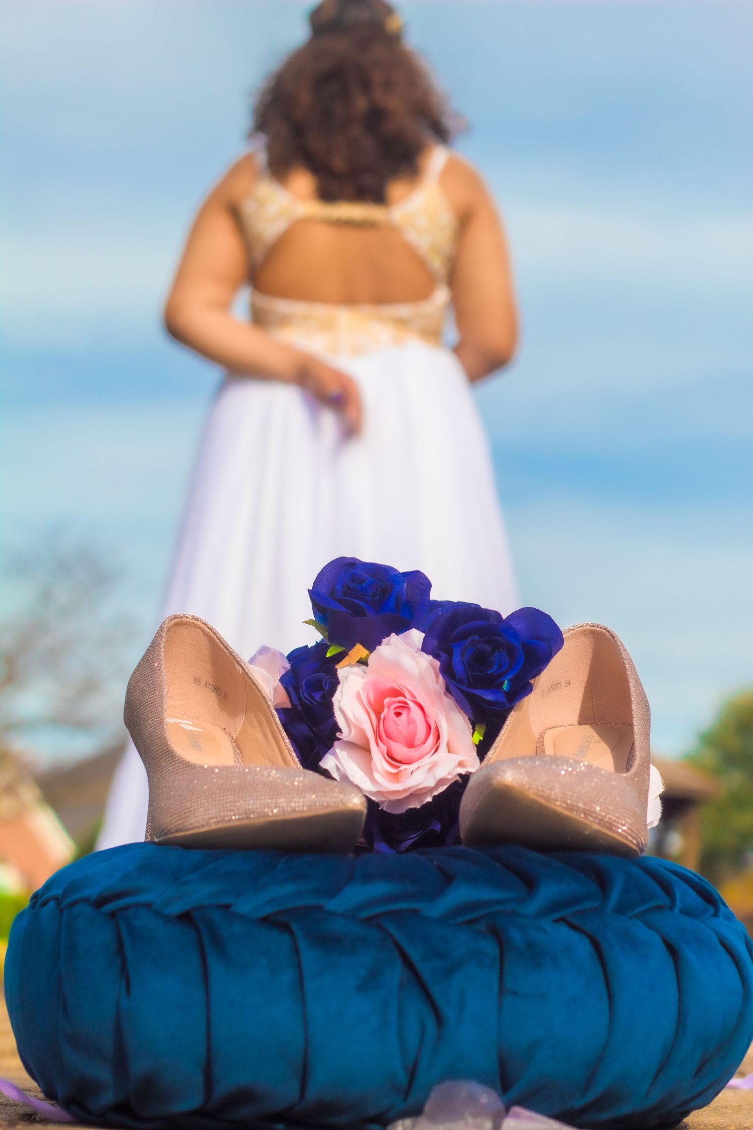 Wedding shoes and roses arranged on blue velvet cushion with bride standing in background.