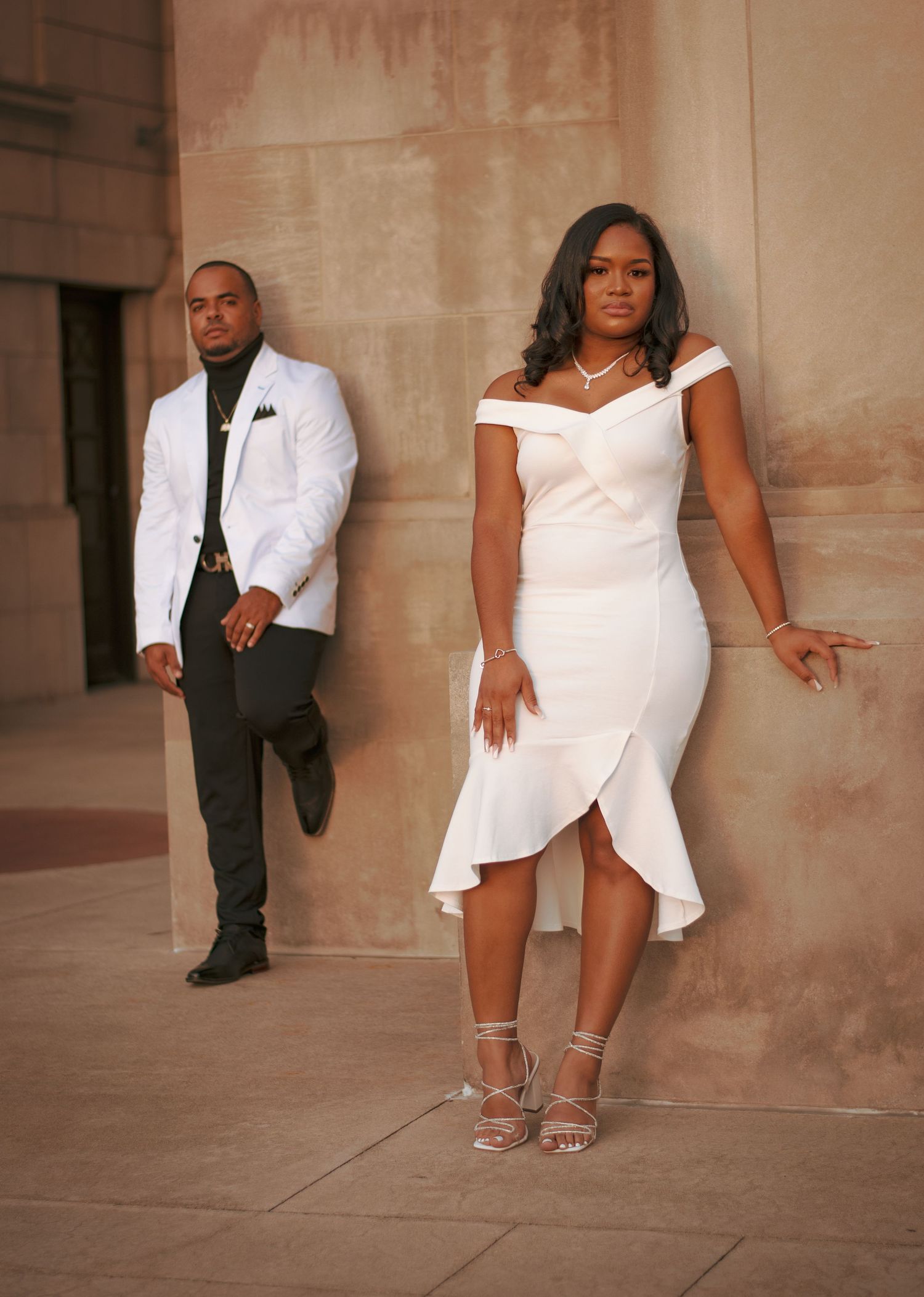 Fashionable portrait of couple in elegant white and black attire against stone wall.