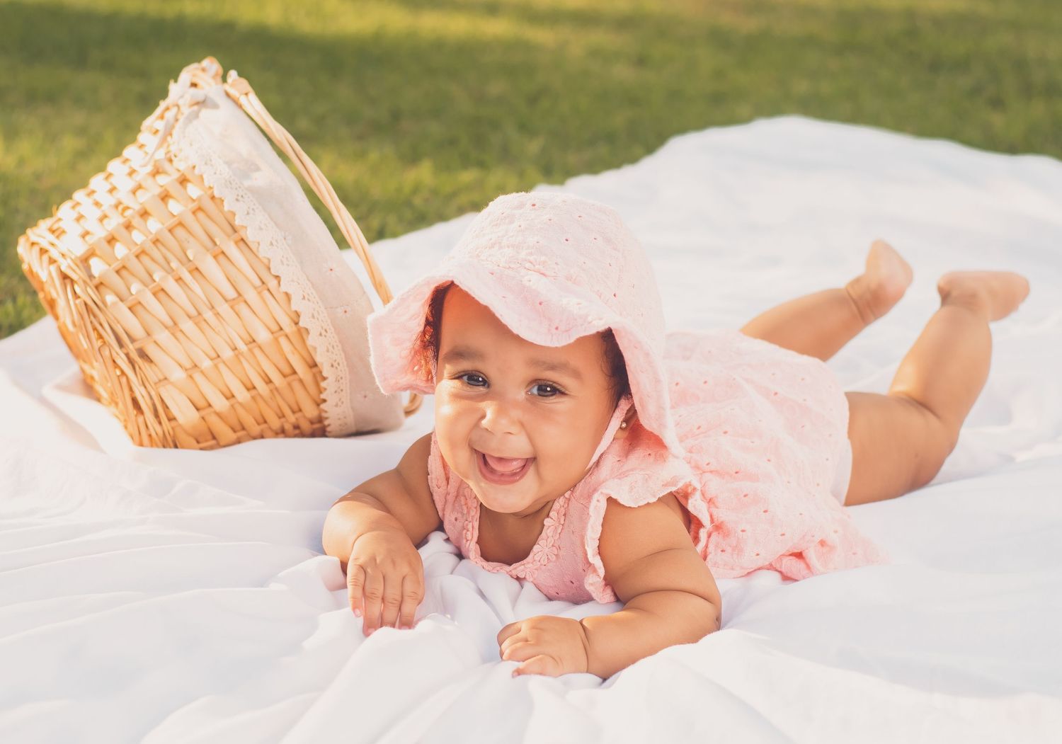 Smiling baby girl in pink outfit and bonnet lying on white blanket outdoors near wicker basket.