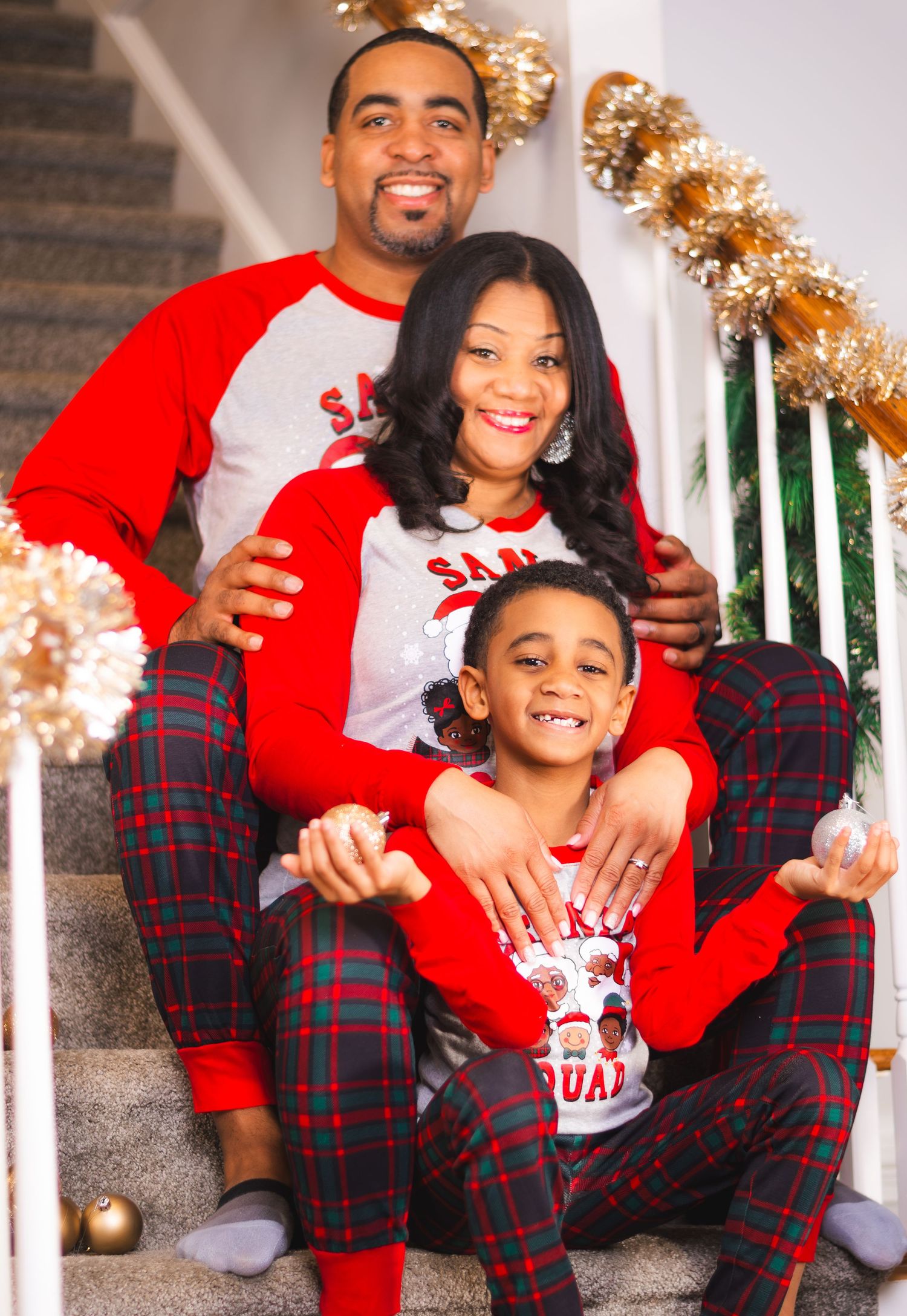 Family of three in matching red plaid Christmas pajamas smiling on staircase decorated with gold holiday garland.
