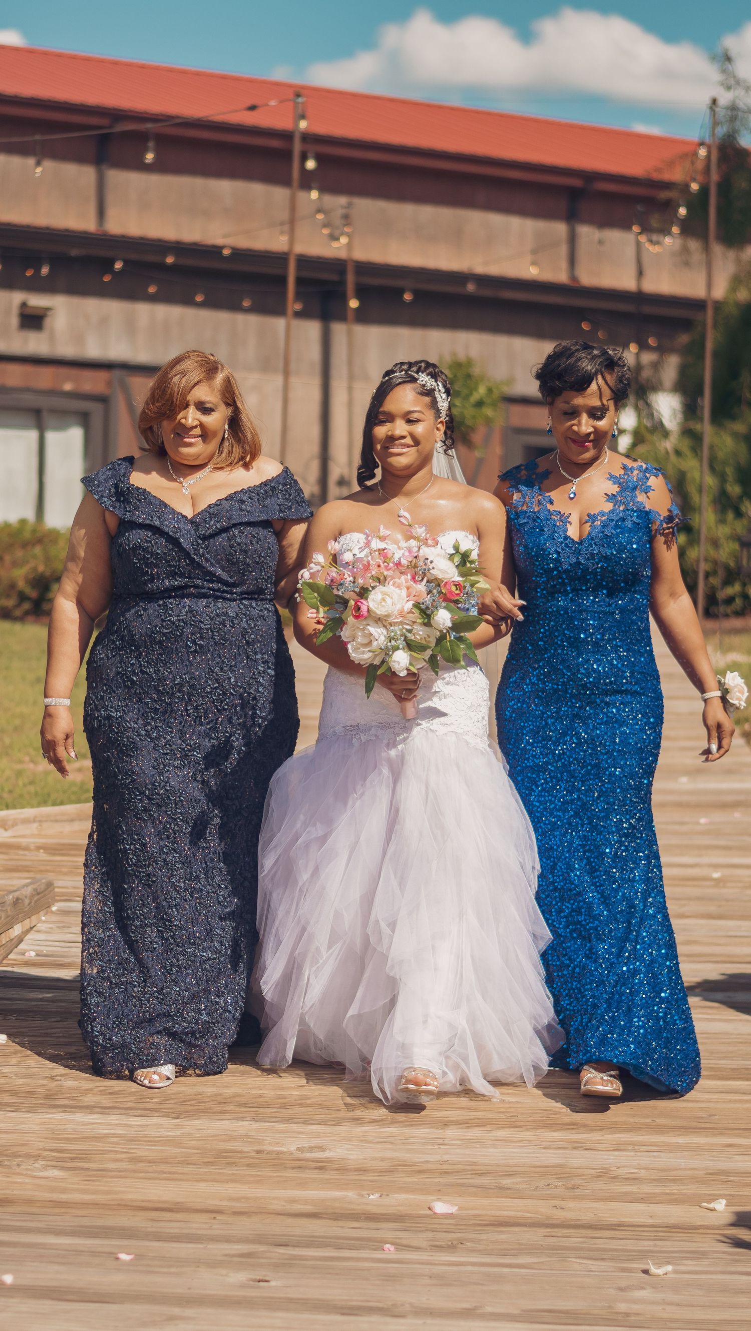 Three women in formal gowns stand together on a wooden deck, with the bride in white holding a pink bouquet.