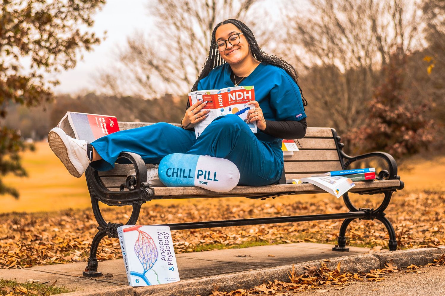 Woman in blue scrubs relaxing on park bench with Chill Pill pillow and NOH magazine among autumn leaves.