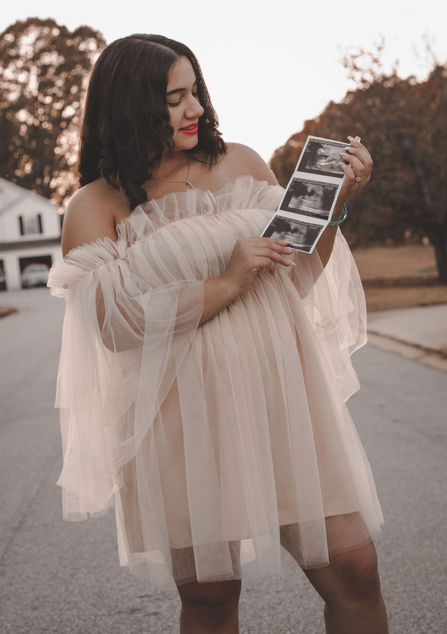Someone wearing a flowy white dress poses with ultrasound pictures on a residential street.