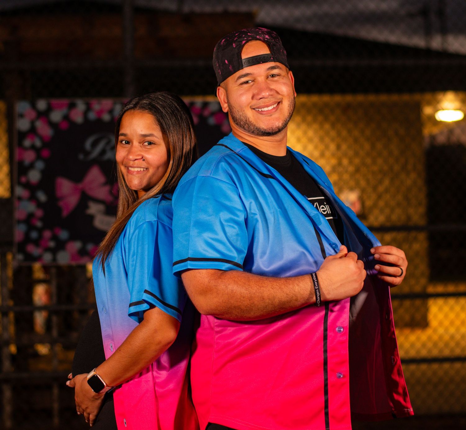 Two people in matching blue and pink gradient jerseys posing back-to-back on a baseball field.