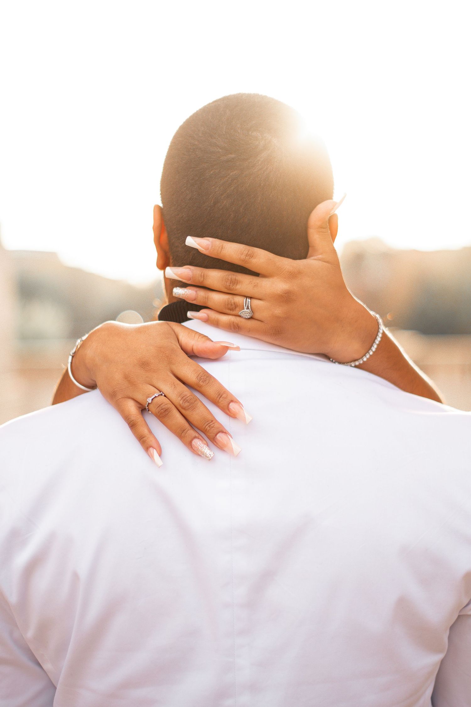 A close-up of hands with engagement rings embracing from behind in warm sunlight.
