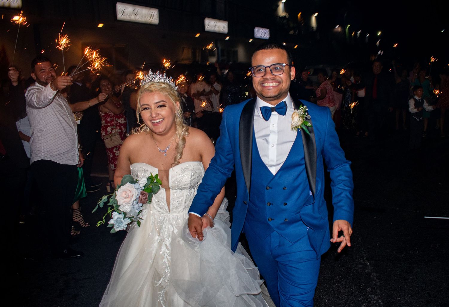 Newlyweds celebrate during nighttime reception with bride in white dress and groom in blue suit.