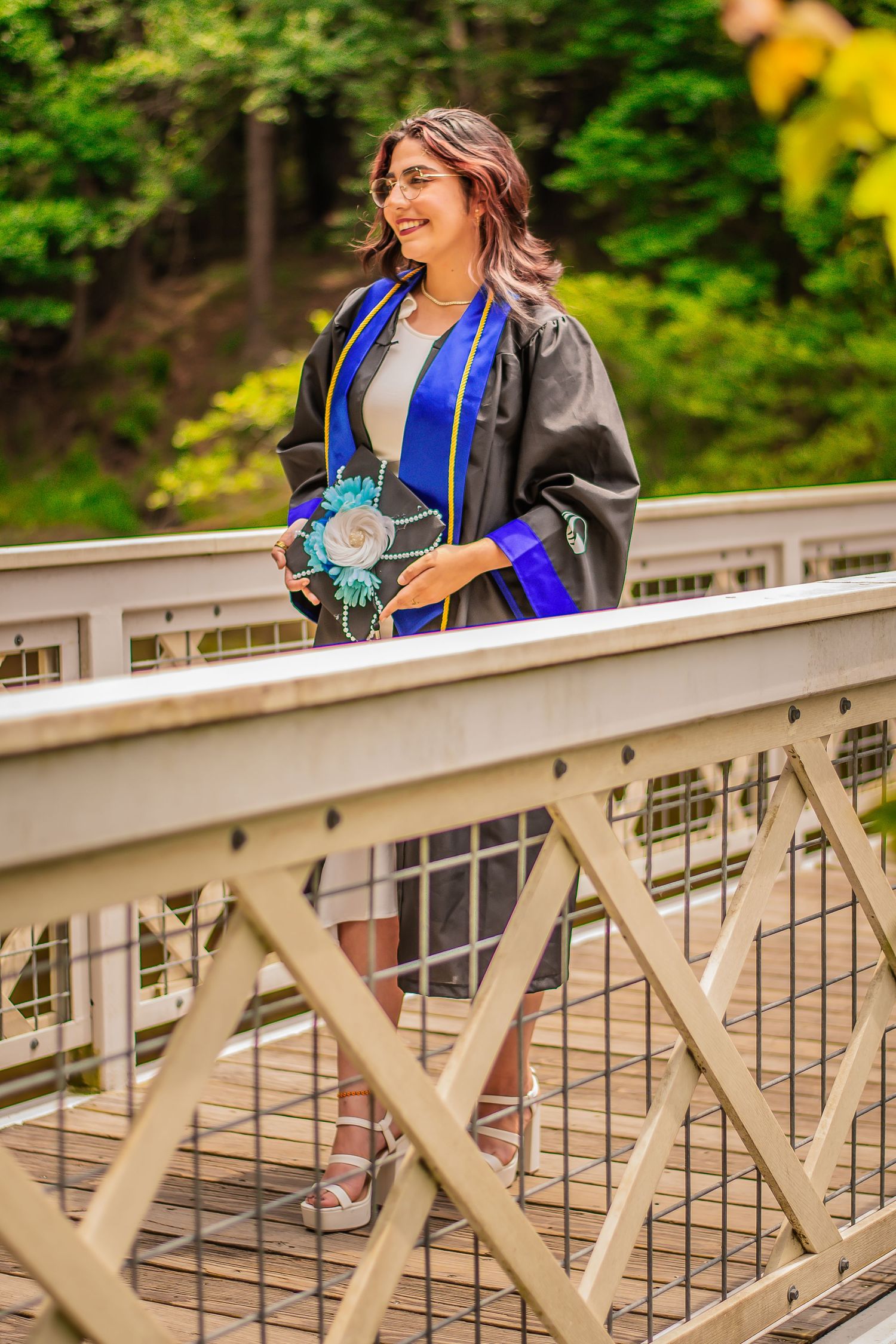 Graduate in blue and black regalia smiling on a bridge in nature.