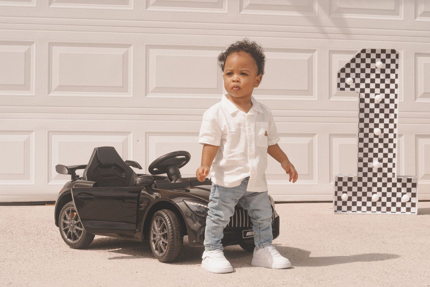 Small child stands next to black ride-on toy car with checkered number one decoration against white garage door.
