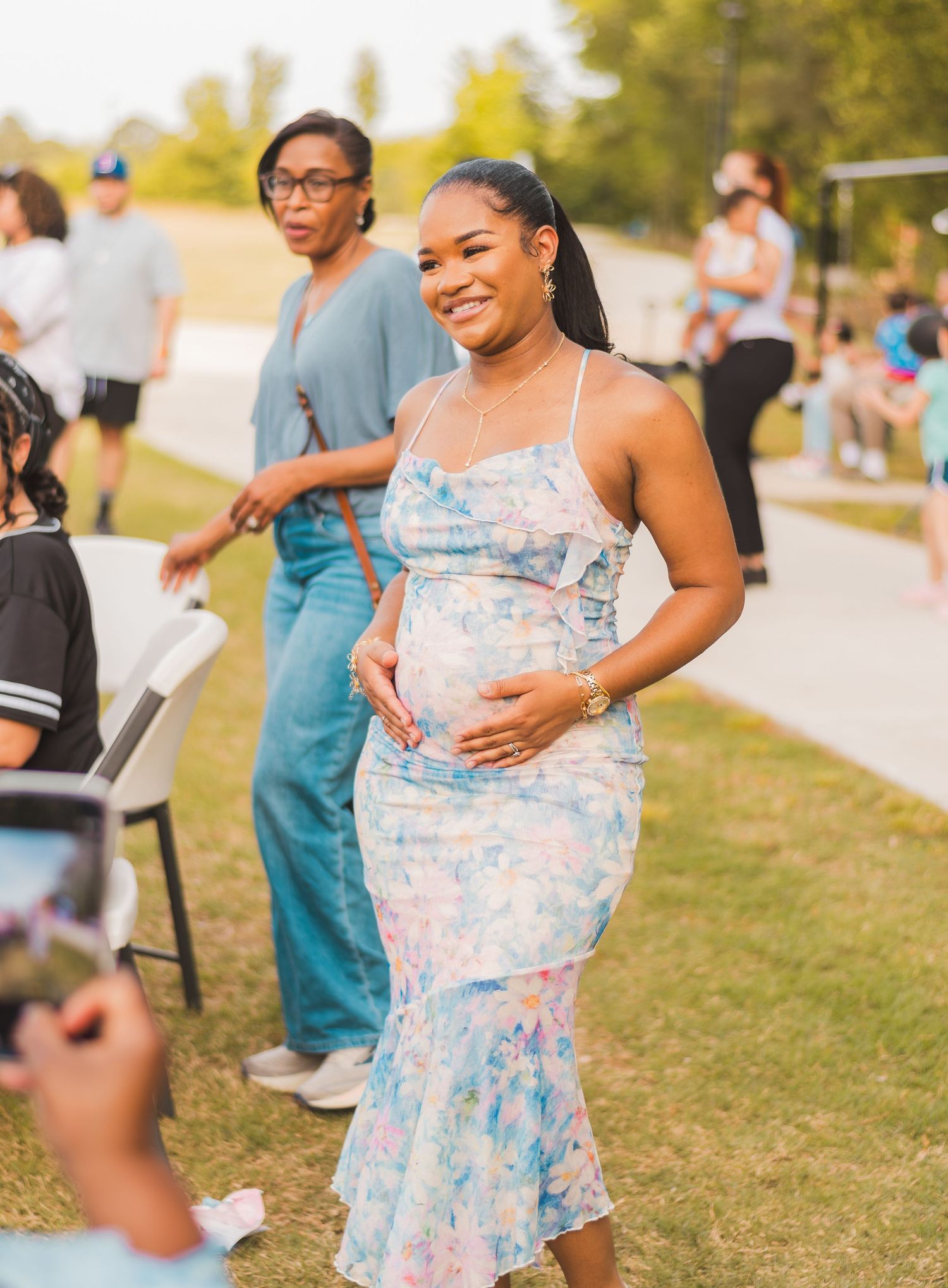 Pregnant woman in floral dress smiling at outdoor event, cradling baby bump.