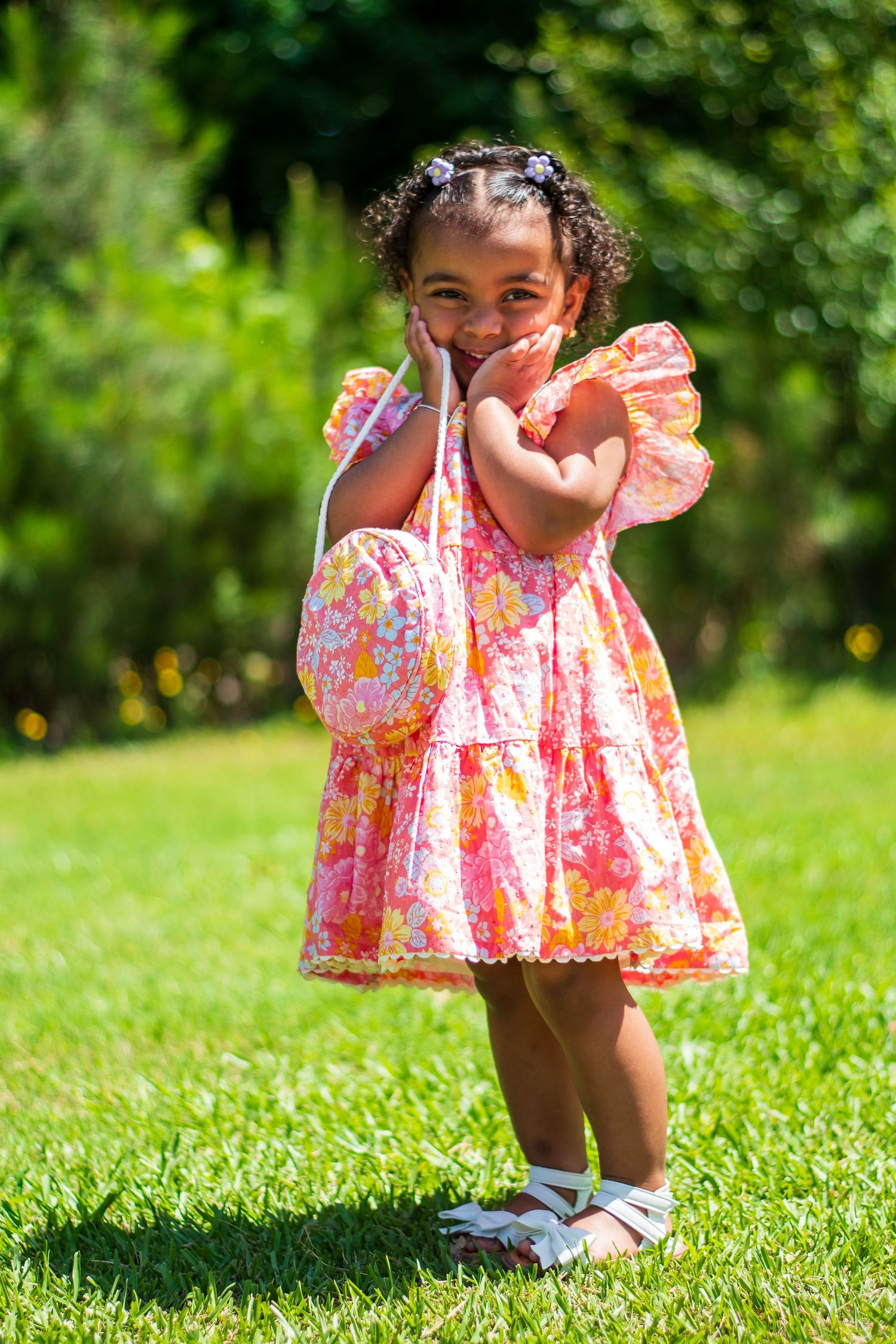 A young child wearing a pink floral dress smiles joyfully while playing outdoors on a sunny day.