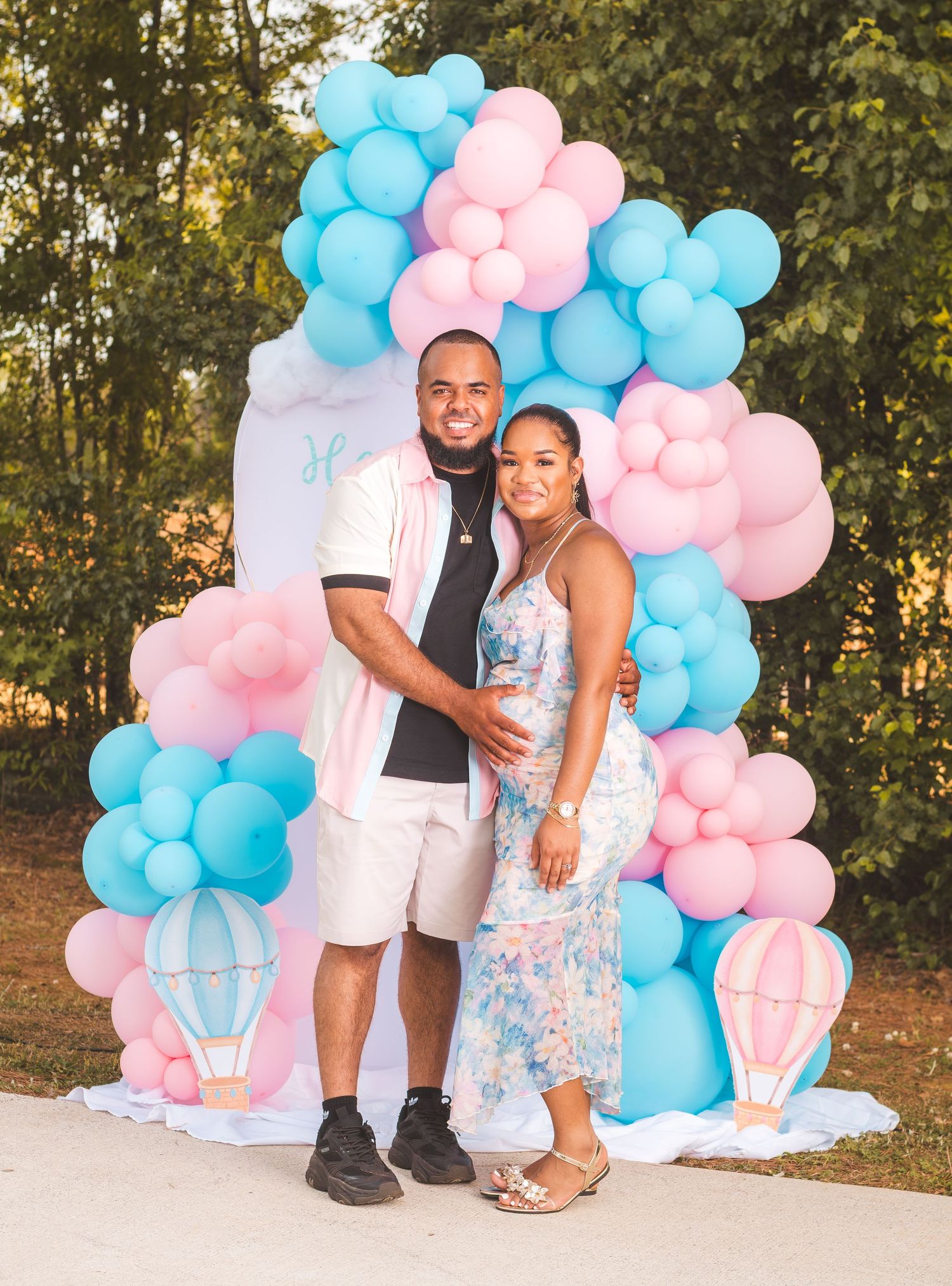 Couple posing at a gender reveal party with pink and blue balloon arch backdrop outdoors.