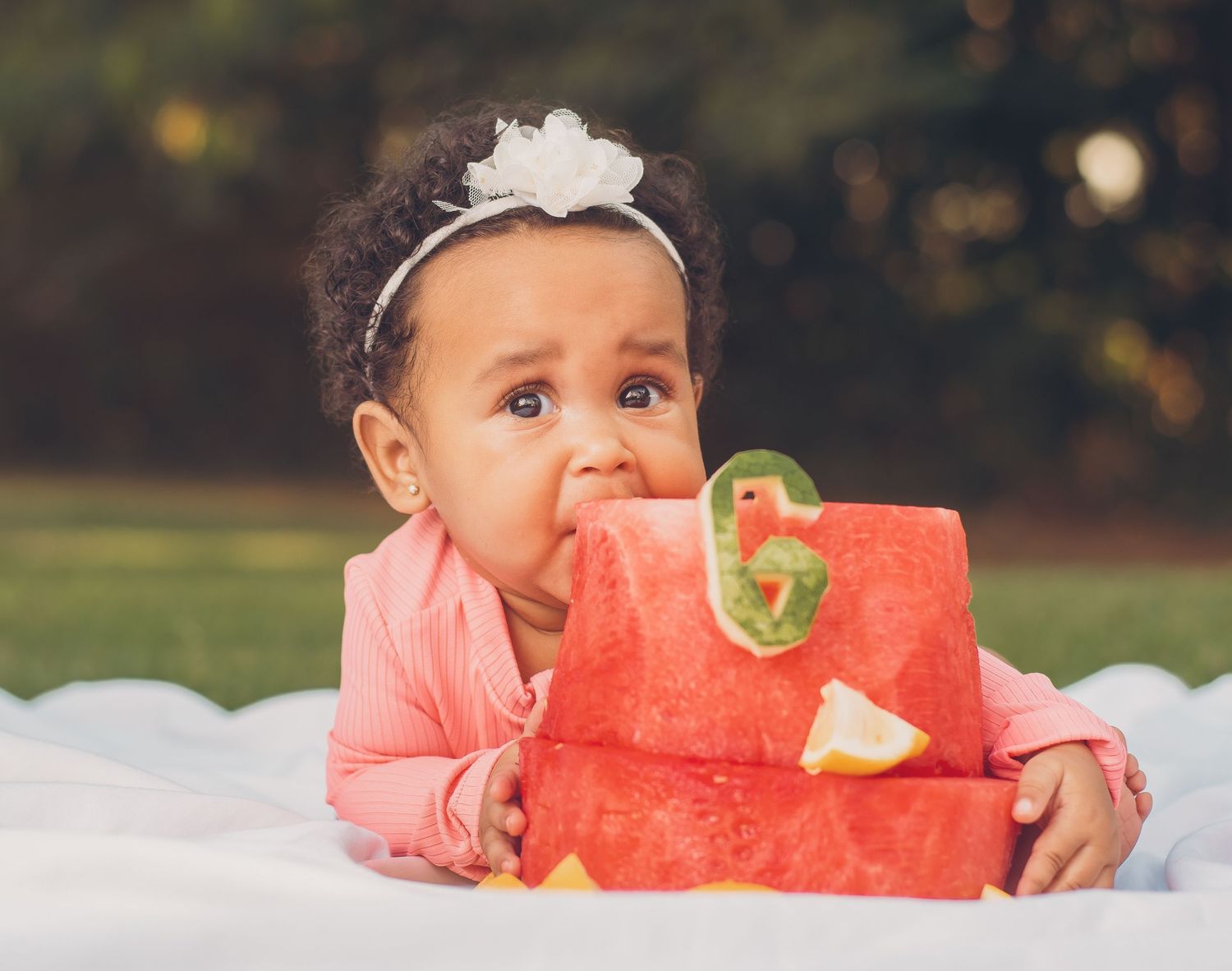 Adorable baby girl in pink outfit eating a watermelon number 6 cake outdoors on a white blanket.