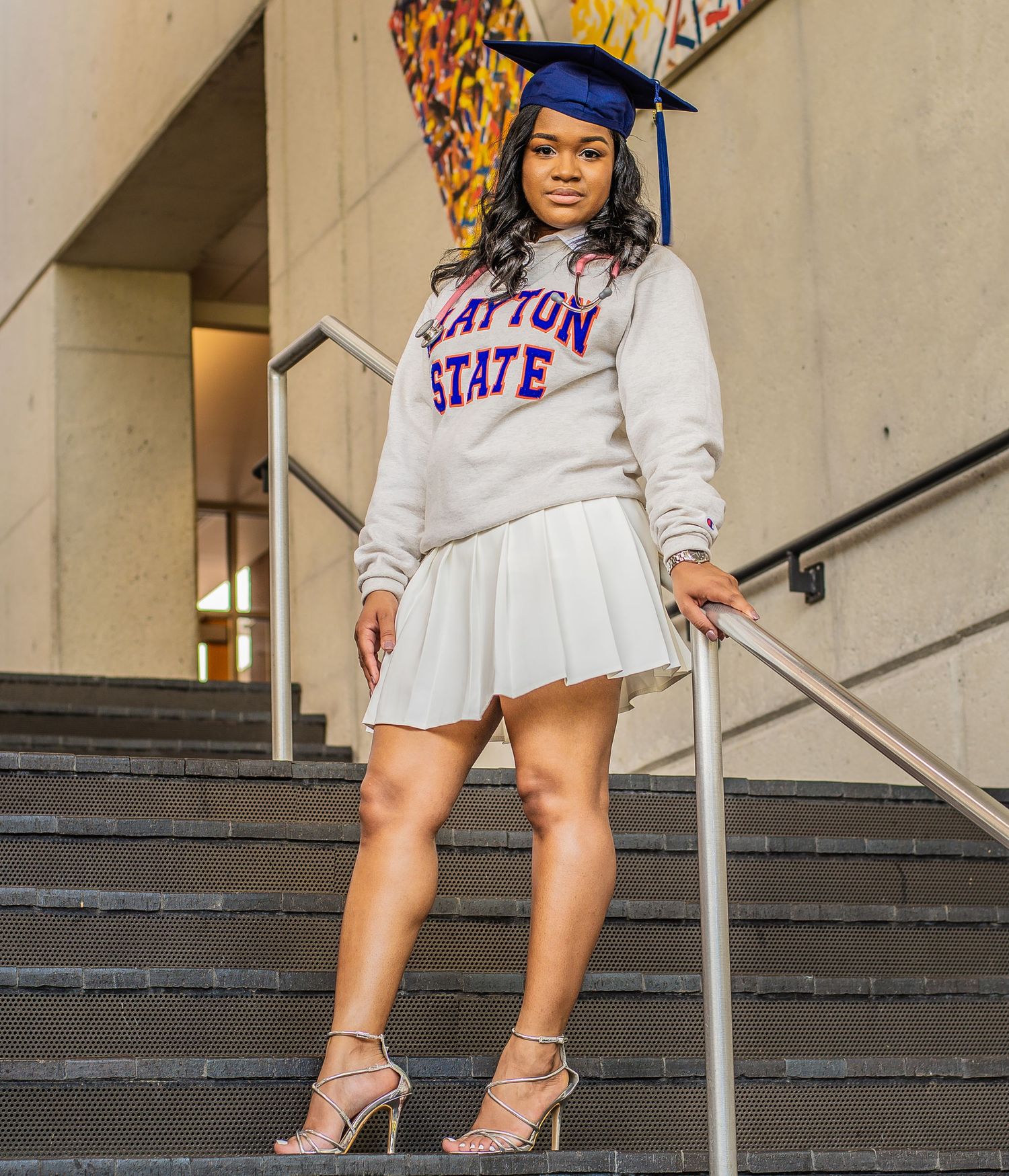 Graduate poses on campus steps wearing university sweatshirt and graduation cap.