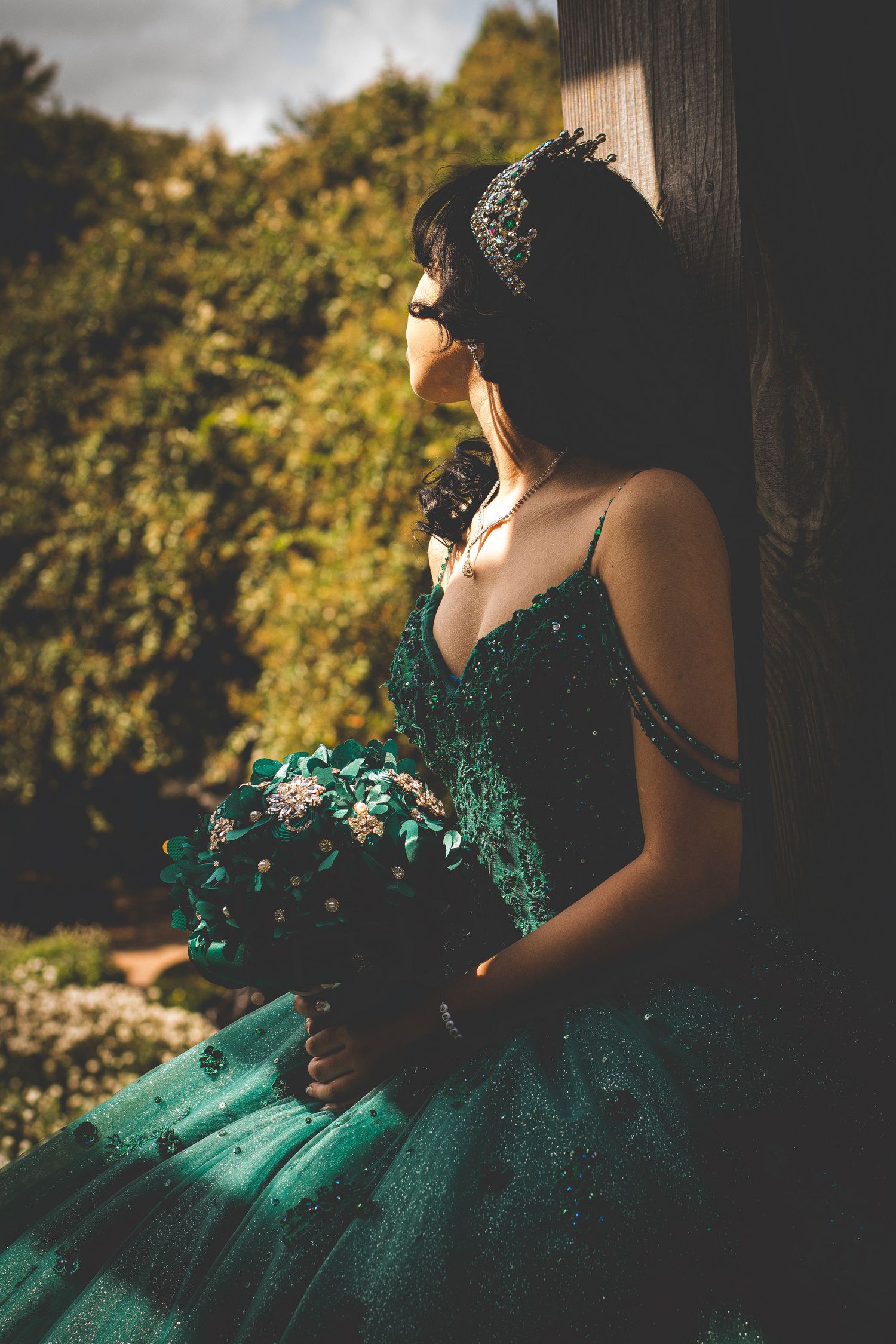 Young woman in emerald green dress with crown holding bouquet in moody outdoor setting.