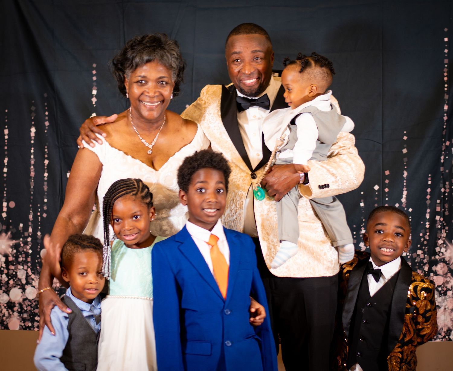 Family portrait in formal attire with sparkly backdrop featuring adults and children in suits.