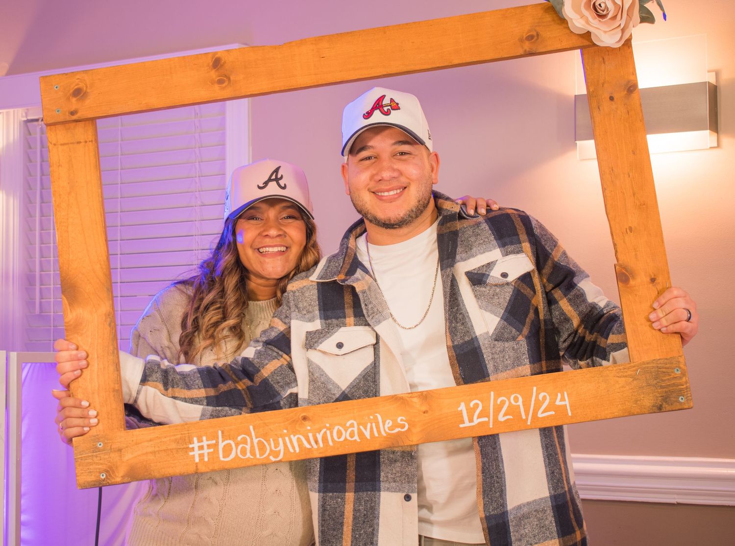 A couple poses with Atlanta Braves baseball caps in a wooden photo frame with the date 12/24/24.