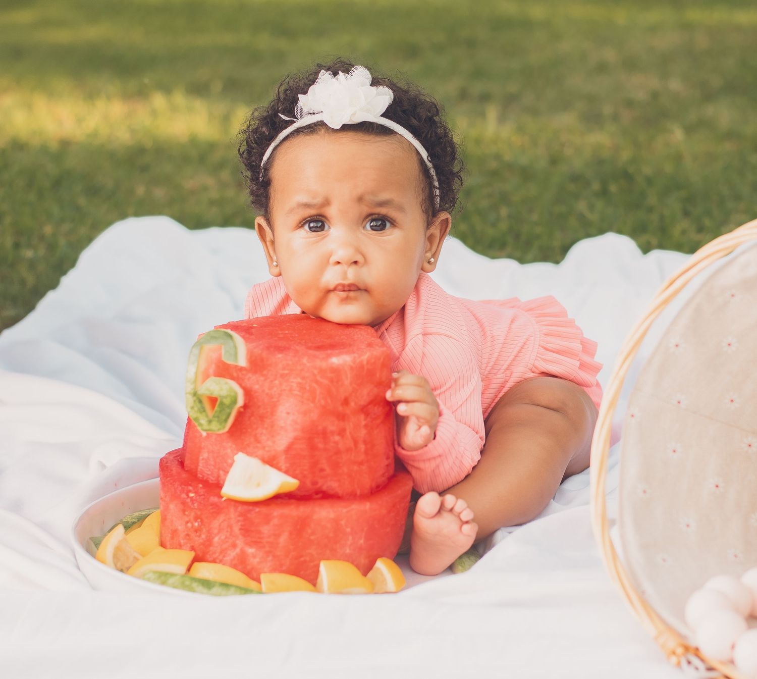 Adorable baby girl in pink outfit biting into a watermelon cake outdoors on a white blanket.