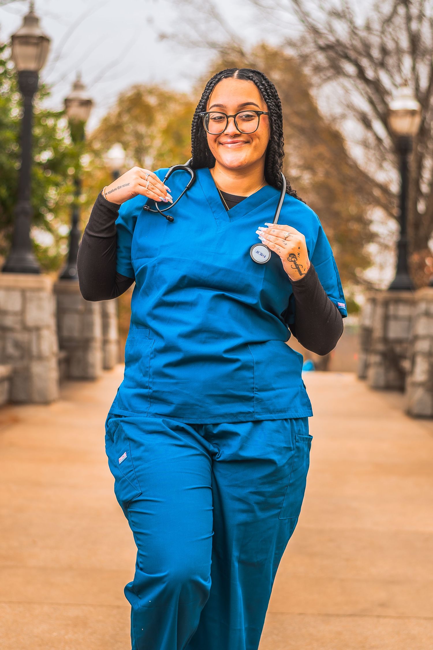 Smiling nurse in blue scrubs holding a stethoscope, walking outdoors on a brick pathway.