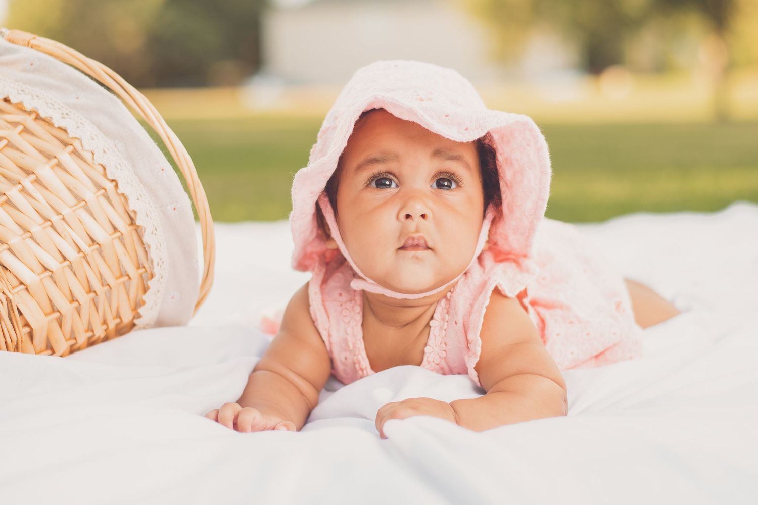 Adorable baby girl in pink bonnet and dress lying on white blanket outdoors near wicker basket.