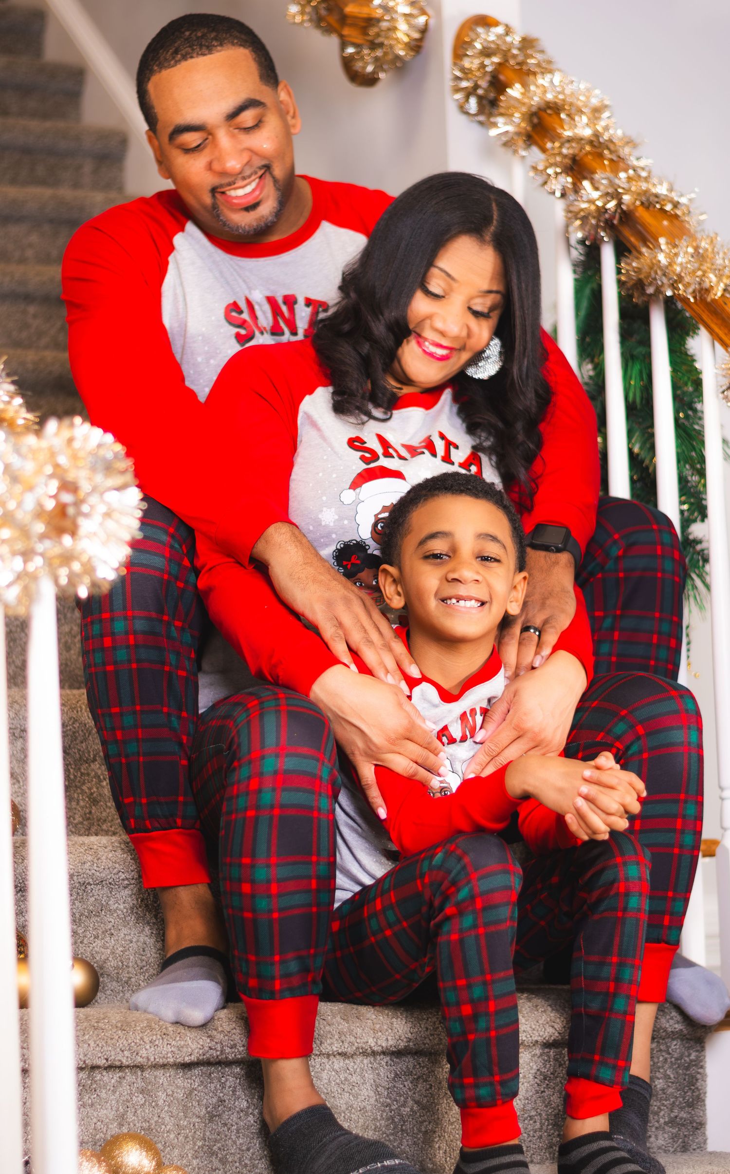 Family of three in matching Christmas plaid pajamas sitting on stairs with holiday decorations.