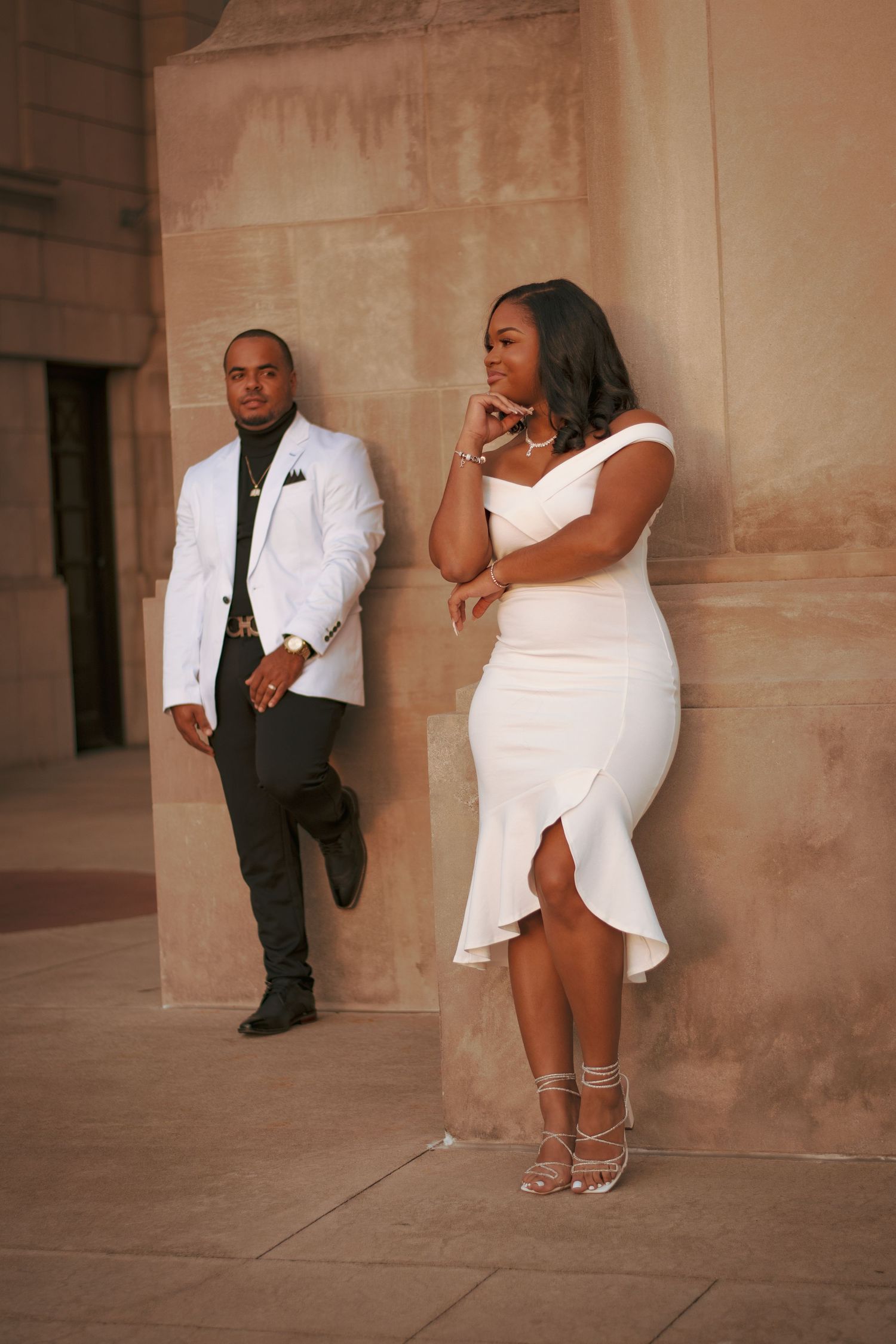 Stylish couple in white and black formal wear posing against beige wall.