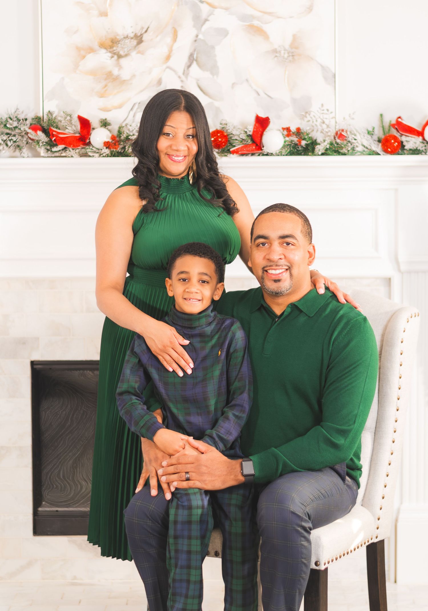 A family of three poses for a Christmas portrait in coordinating green outfits near a festive holiday fireplace mantel.