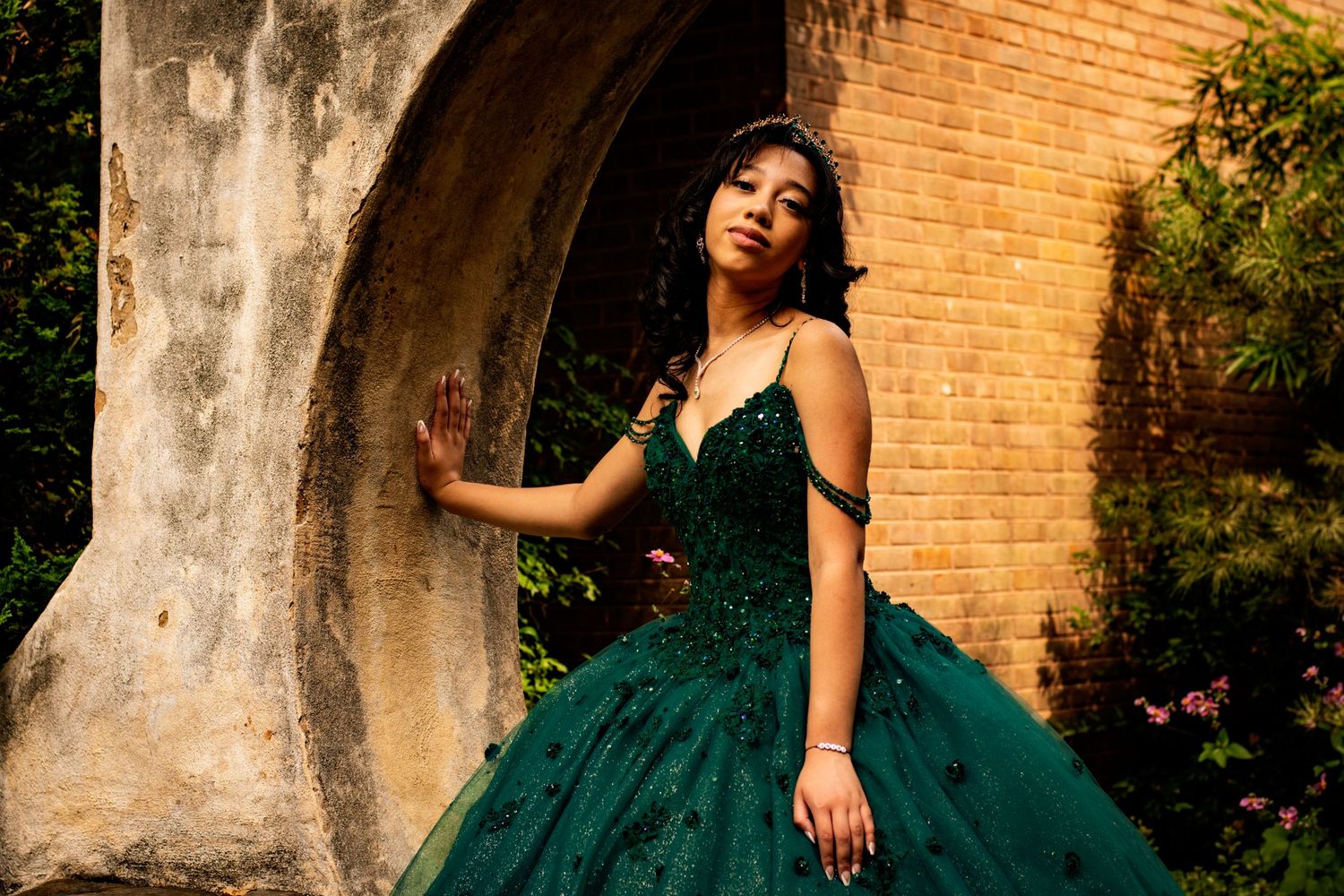 A stunning emerald green ball gown captured against a rustic brick wall and garden backdrop.