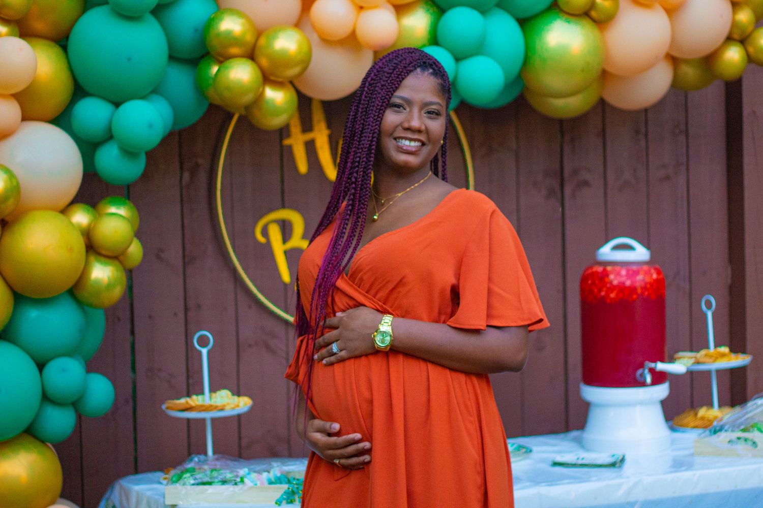 Expecting mother in orange dress poses at baby shower with decorative balloon display.