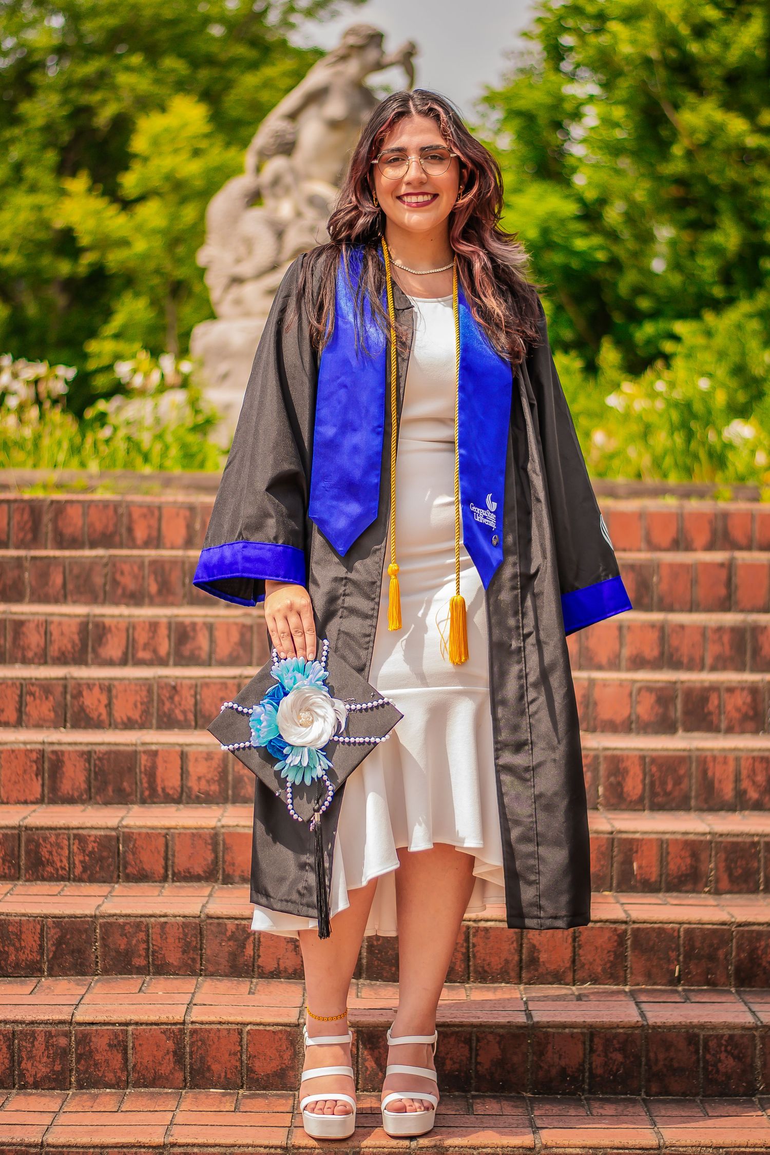 Graduate in black gown with blue stole posing on brick stairs outdoors.