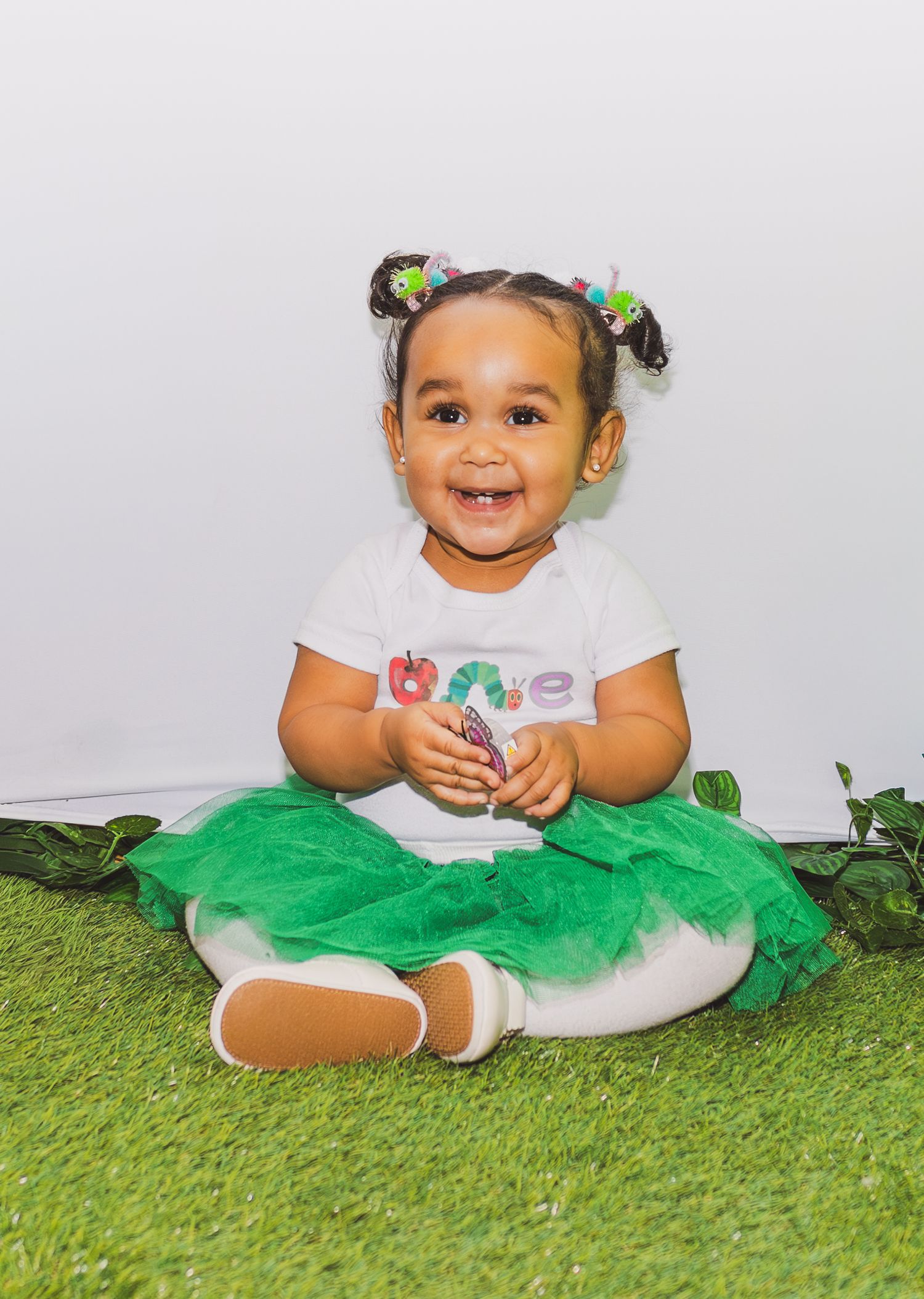 Smiling toddler girl in green tutu and white shirt sits on grass for a first birthday photo shoot.
